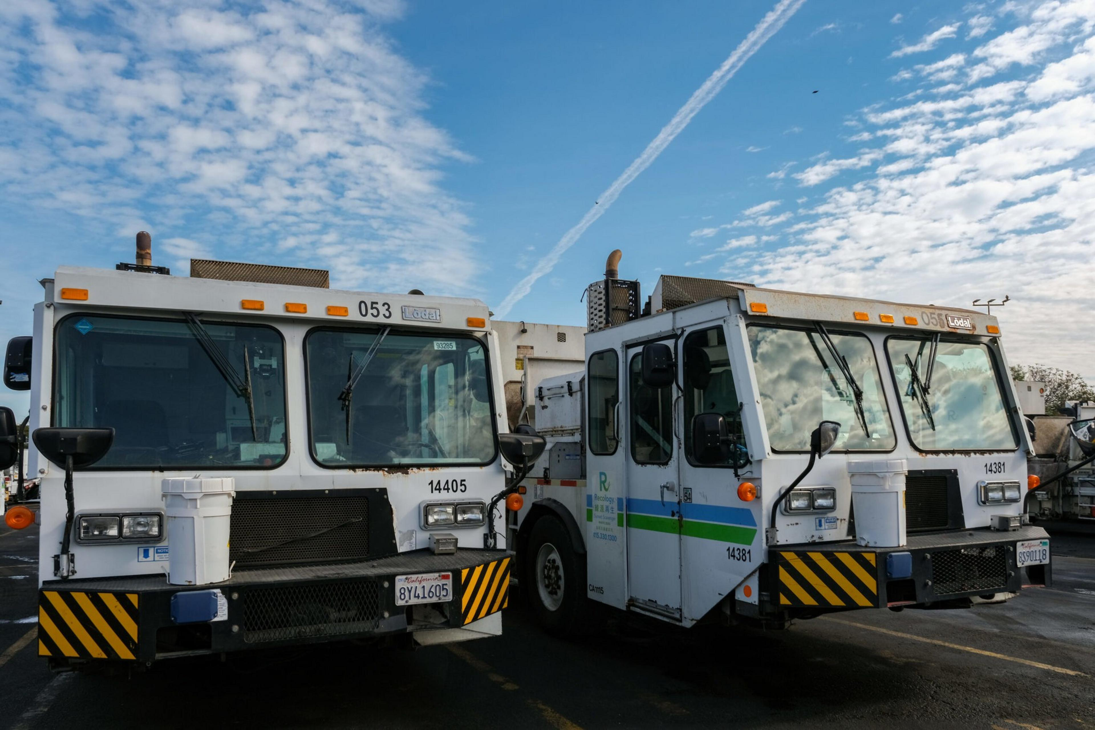 Two white garbage trucks with black and yellow striped bumpers are parked side by side under a blue sky with scattered clouds.