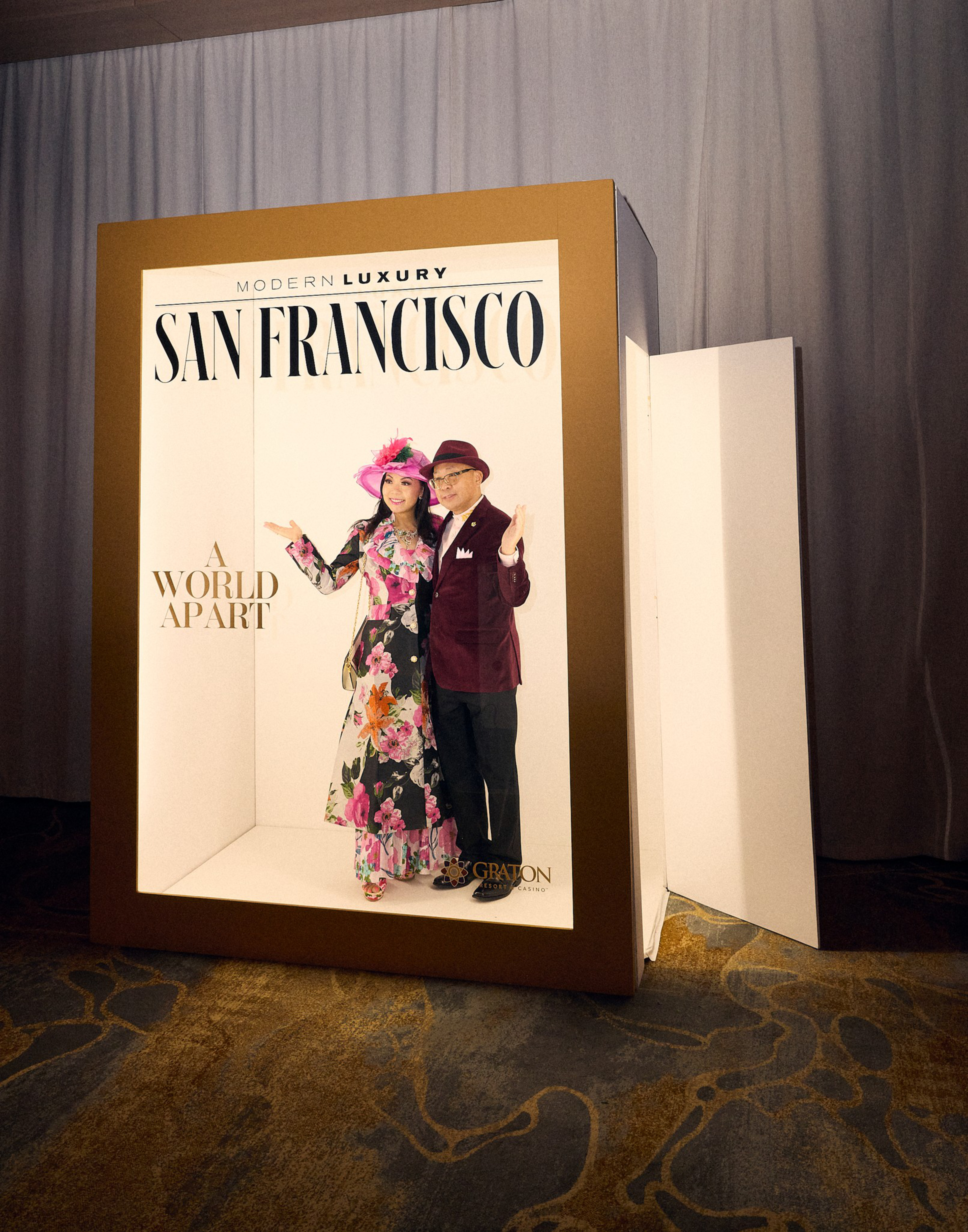 A woman in a floral dress with a pink hat and a man in a maroon suit and hat pose inside a large box designed like a “San Francisco” magazine cover.