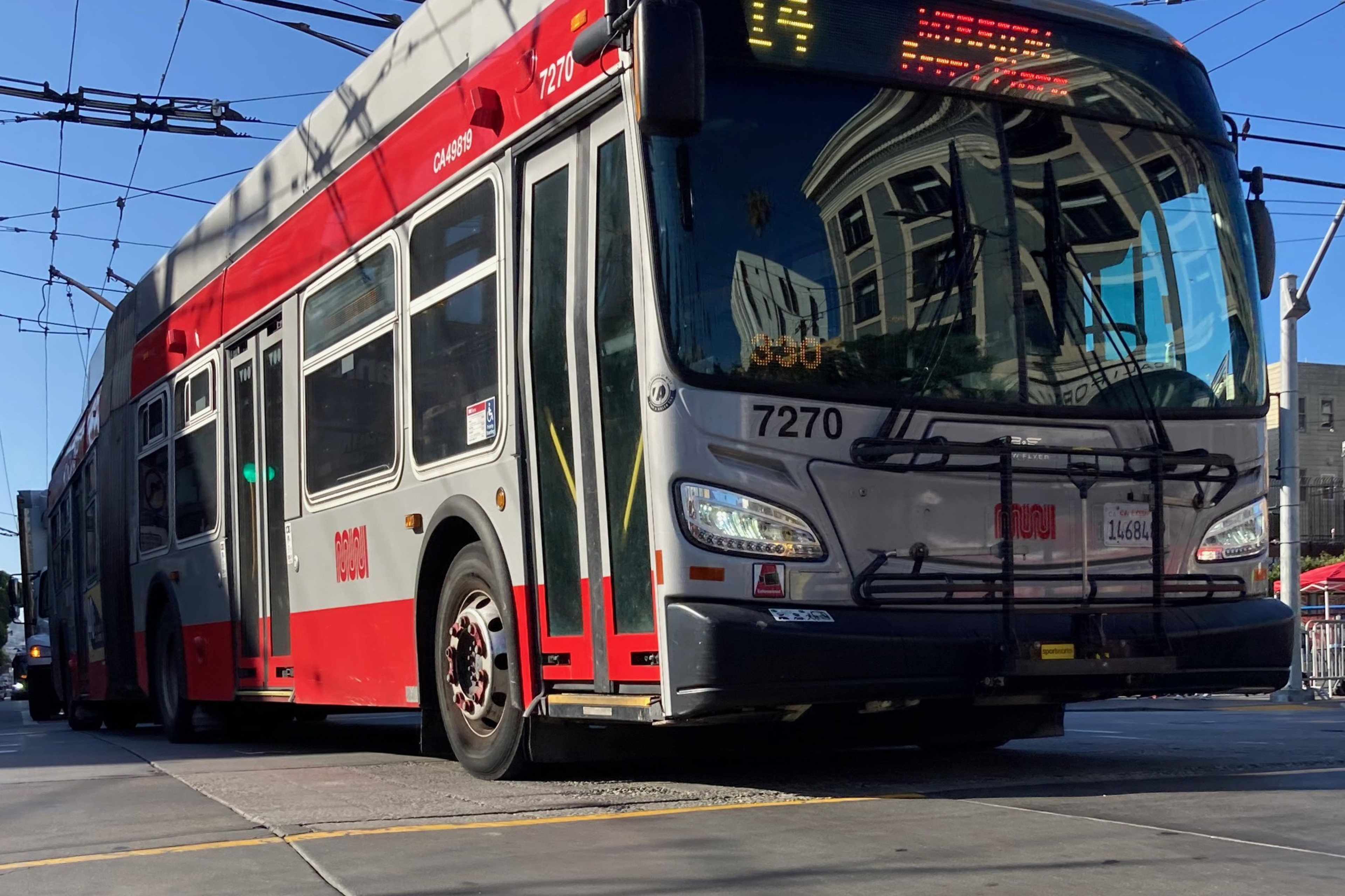 A red and gray city bus is driving on a street, with overhead wires visible above. It has the number 7270 on the front and a building reflection on its windshield.