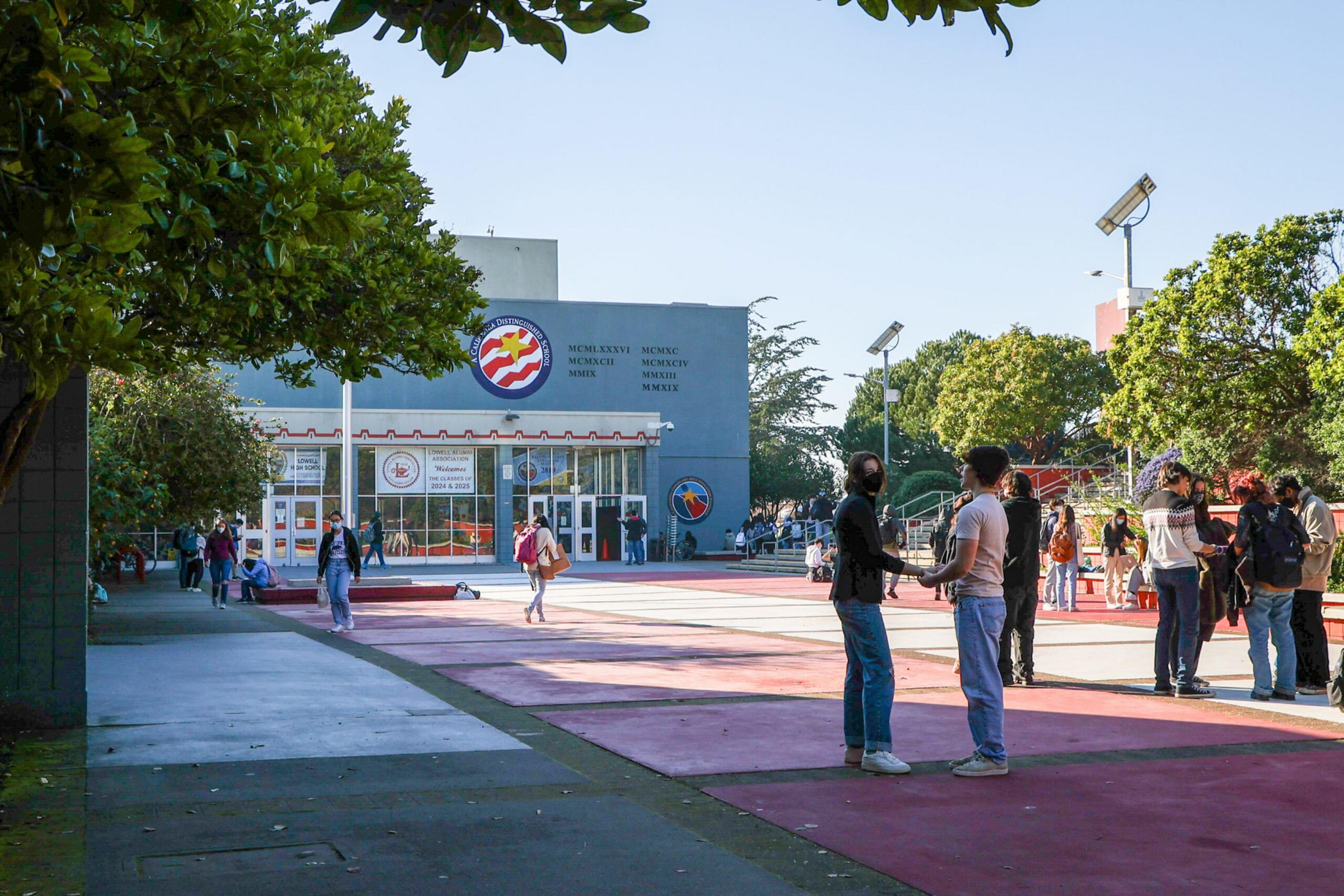 Students wearing masks walk, talk, and stand in small groups outside a school building under a clear blue sky.