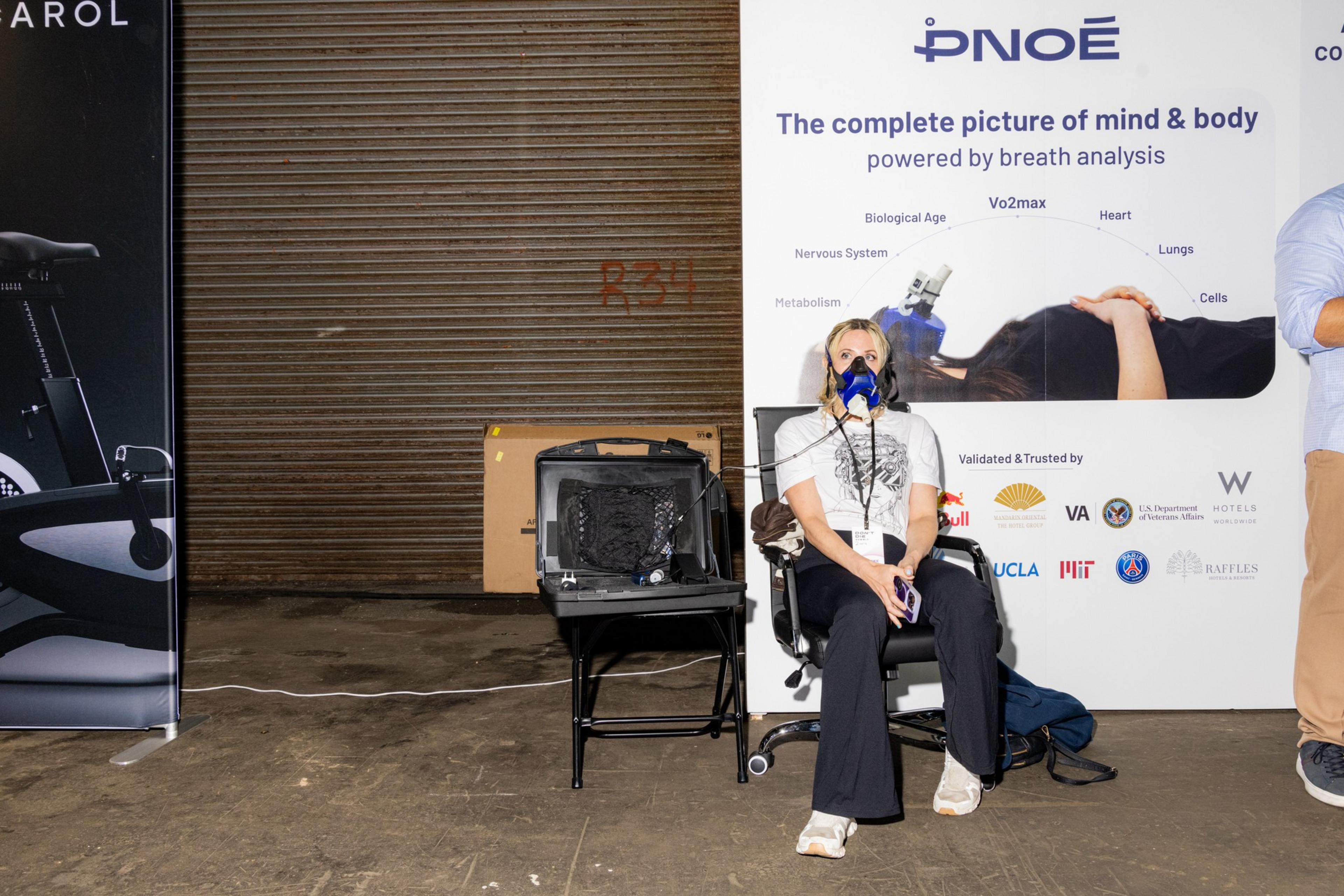 A person sits in a chair wearing a breath analysis mask, next to an open case with equipment and a backdrop displaying &quot;PNOÉ&quot; and health analysis details.