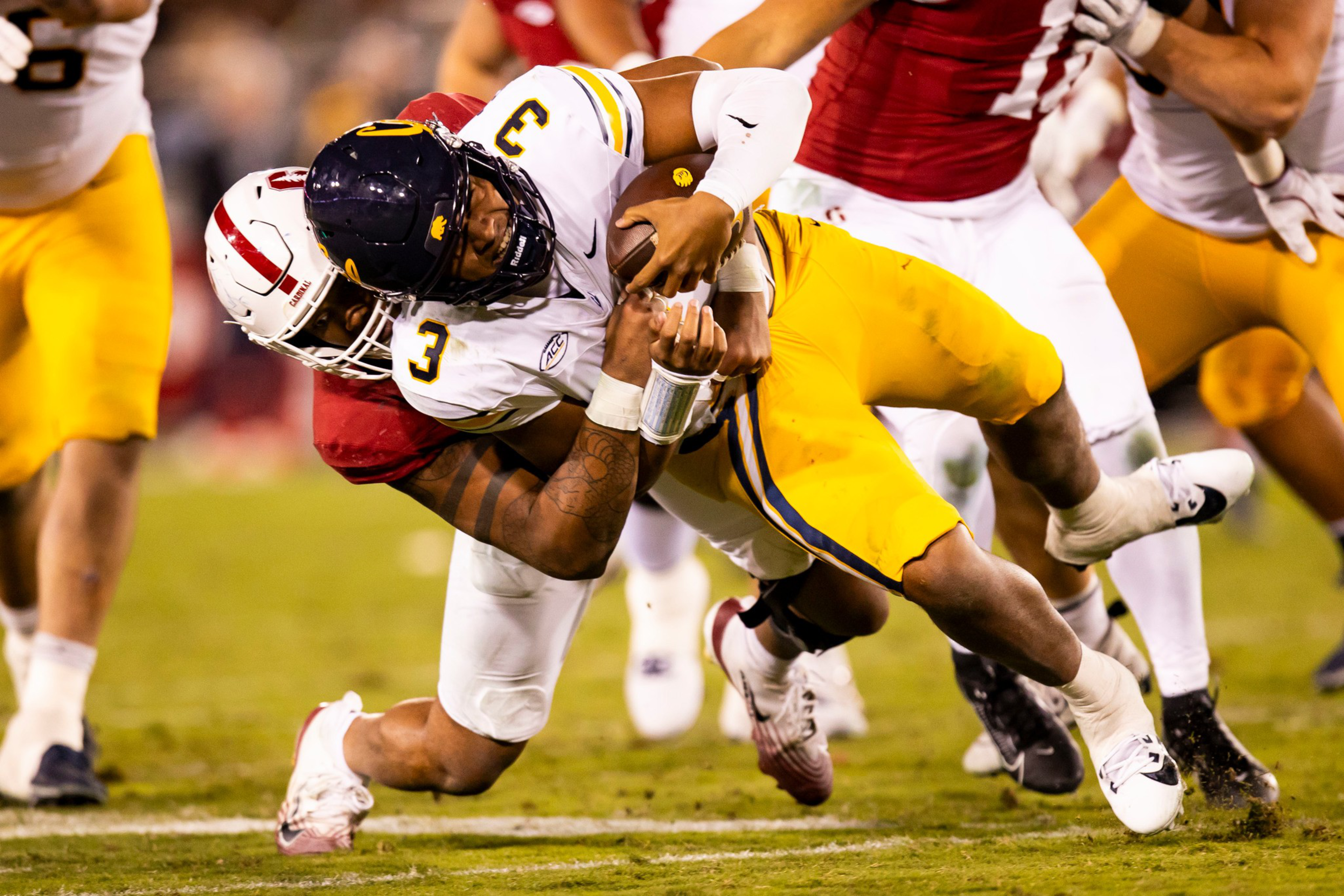 A football player in a white jersey with yellow pants is tackled mid-air by a player in a red jersey during an intense game.