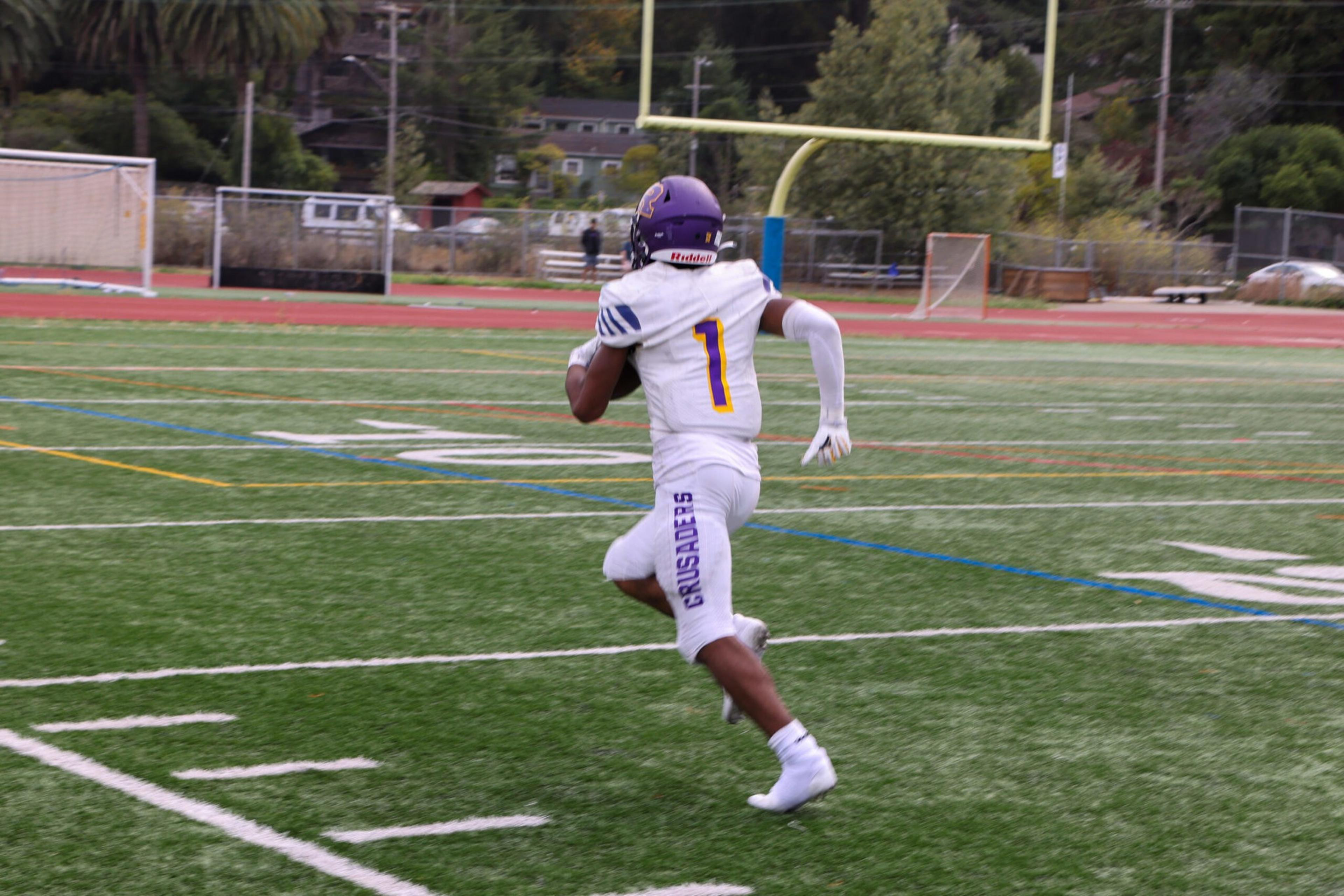 A football player wearing a white uniform with the number 1 and "Crusaders" runs on a green field toward the goalpost holding a football.