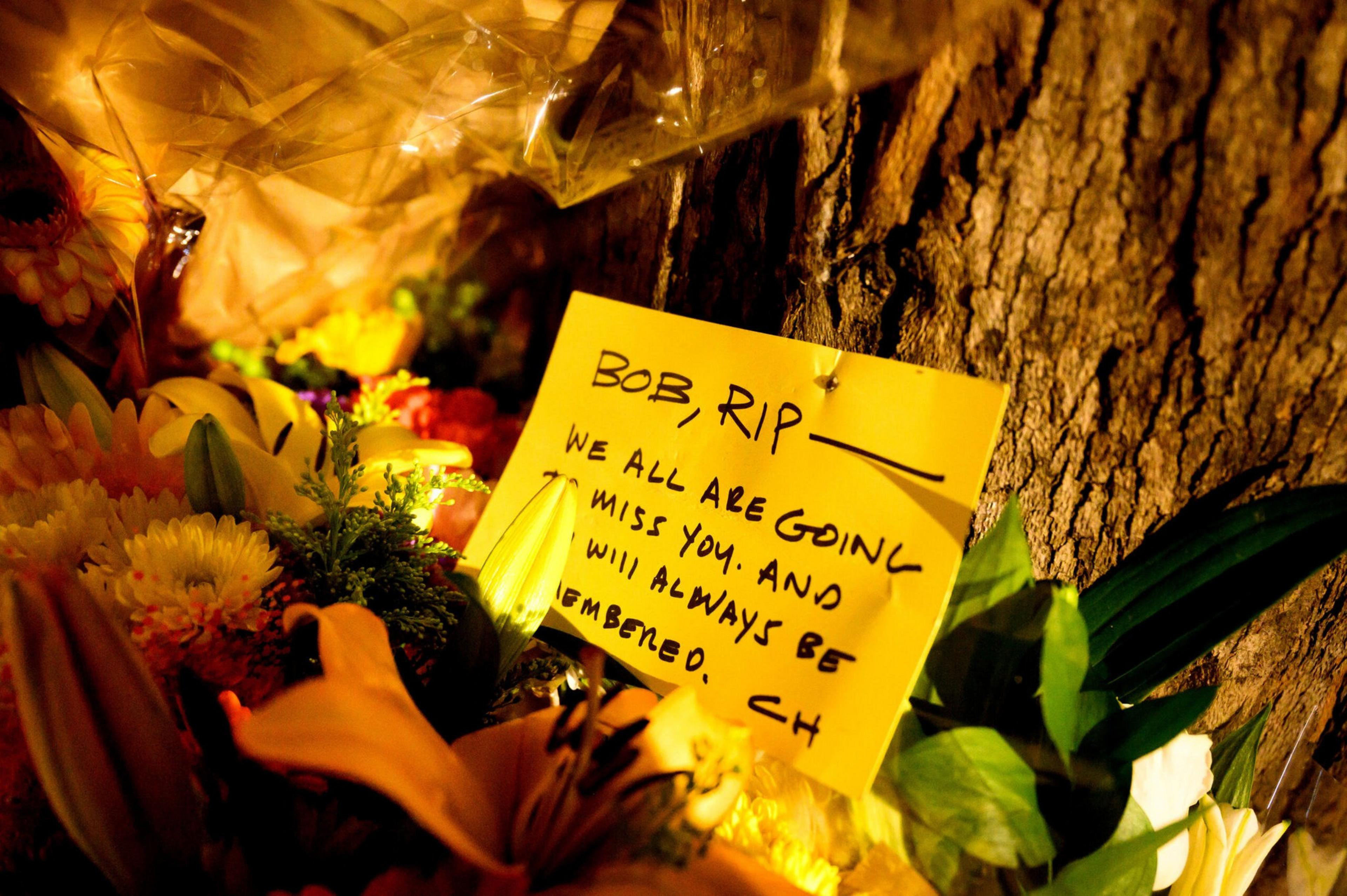 A yellow note pinned to a tree reads "BOB, RIP. We all are going to miss you and will always be remembered," surrounded by colorful flowers.