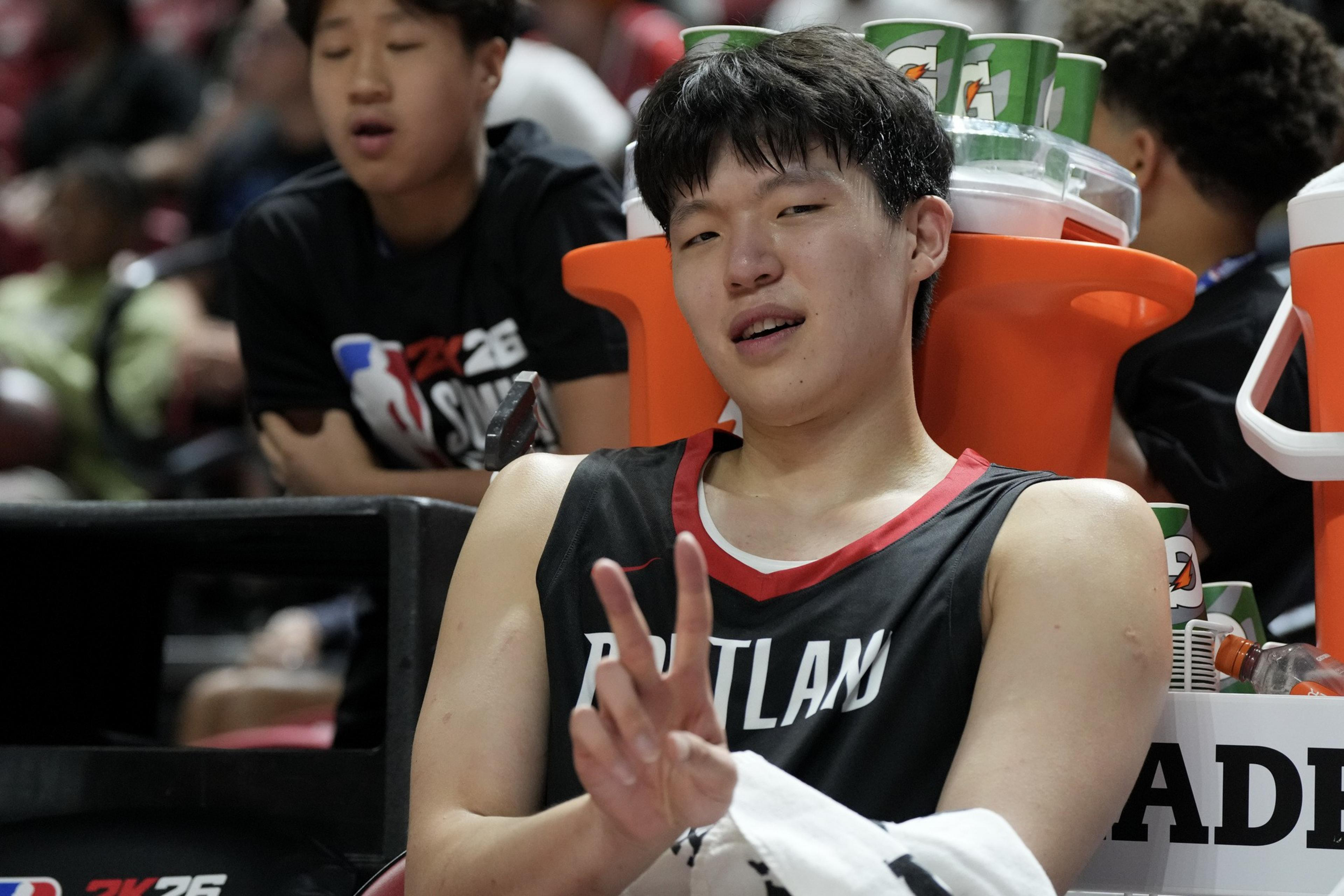 A basketball player in a Portland jersey poses with a peace sign while sitting near Gatorade containers. Another person is in the background, eyes closed.