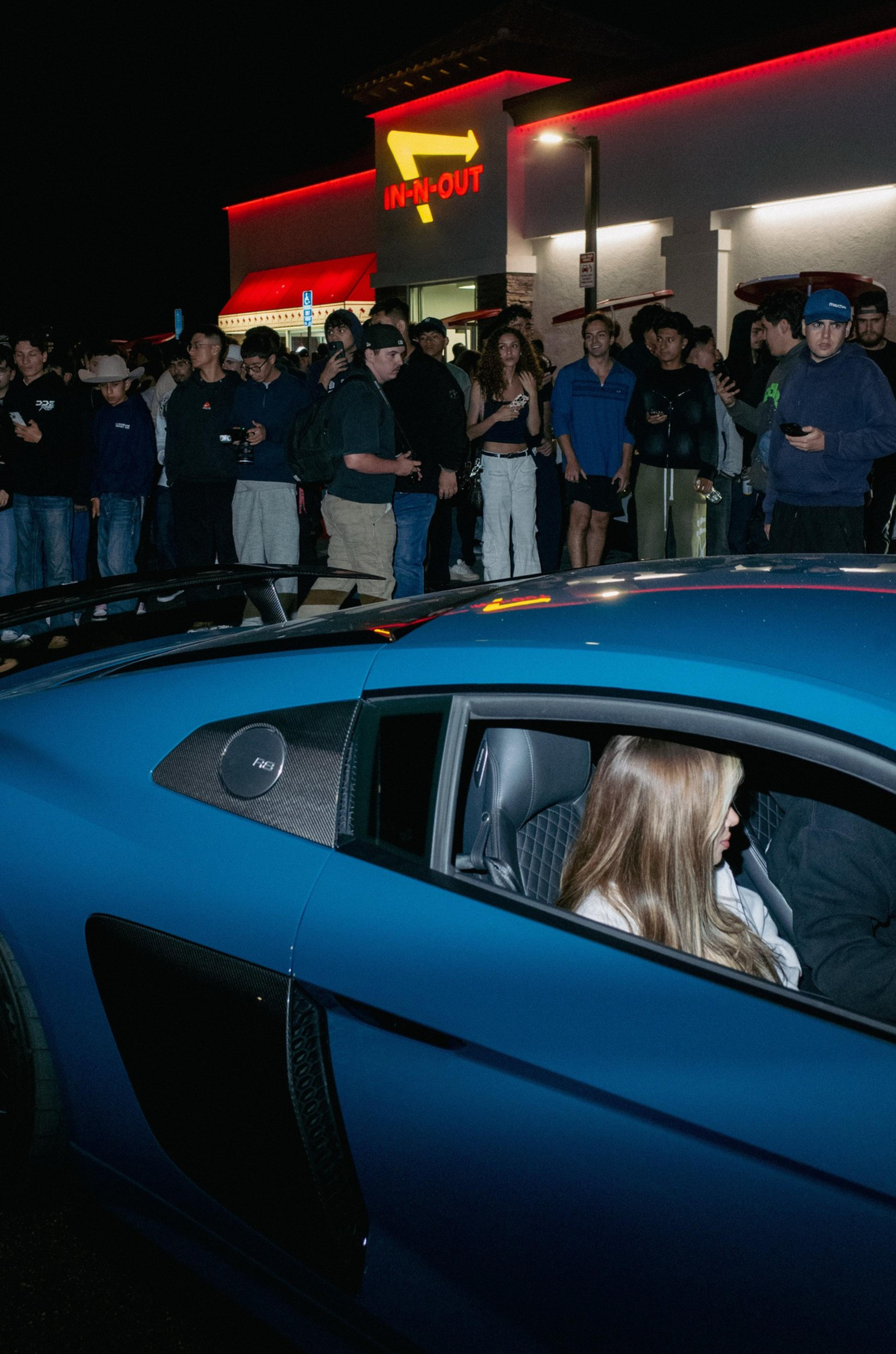 A crowd gathers outside an In-N-Out at night, watching a blue sports car with a woman sitting inside by the driver’s seat.