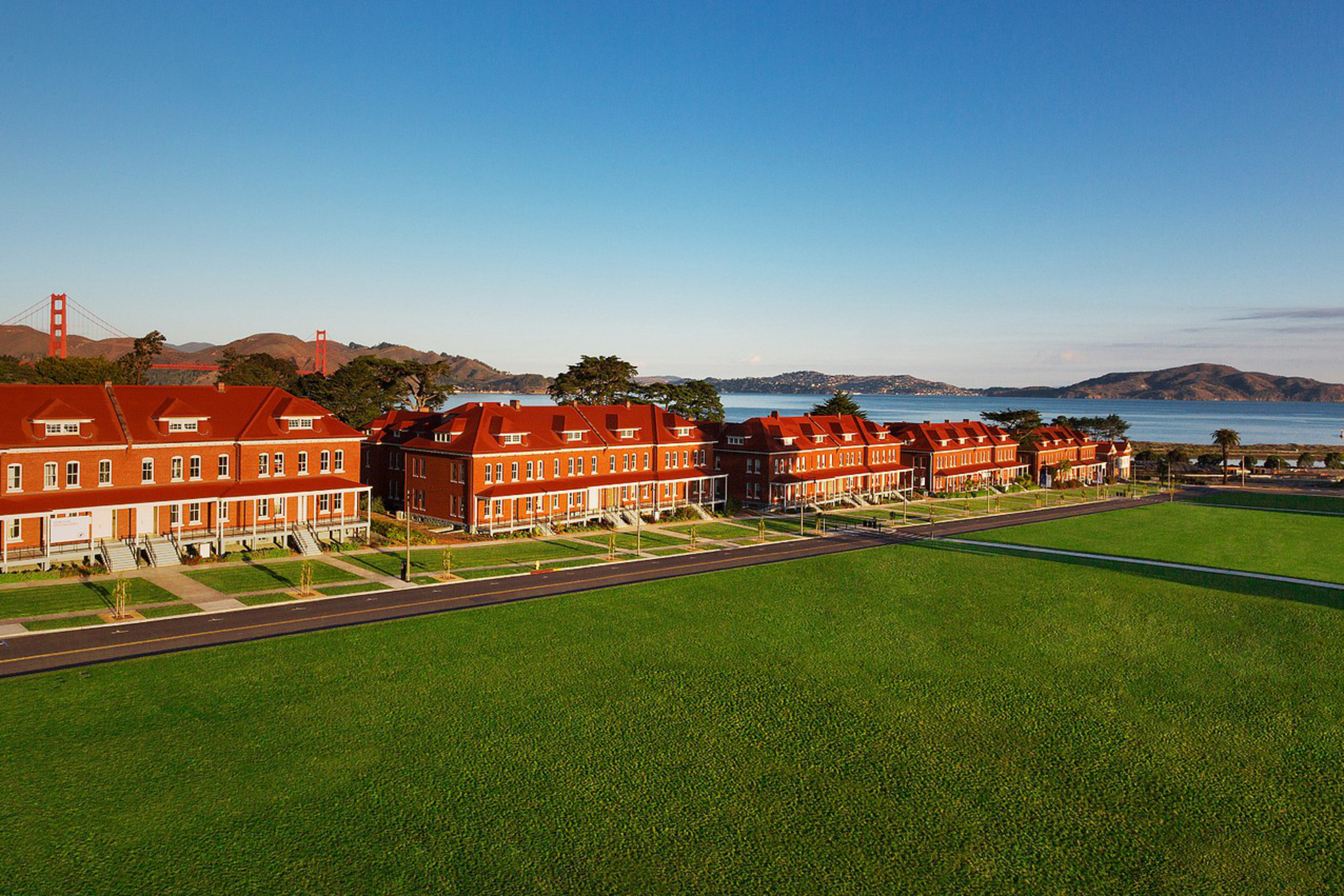 Row of red-roofed buildings with Golden Gate Bridge in the distance, beside a green lawn under clear blue skies.