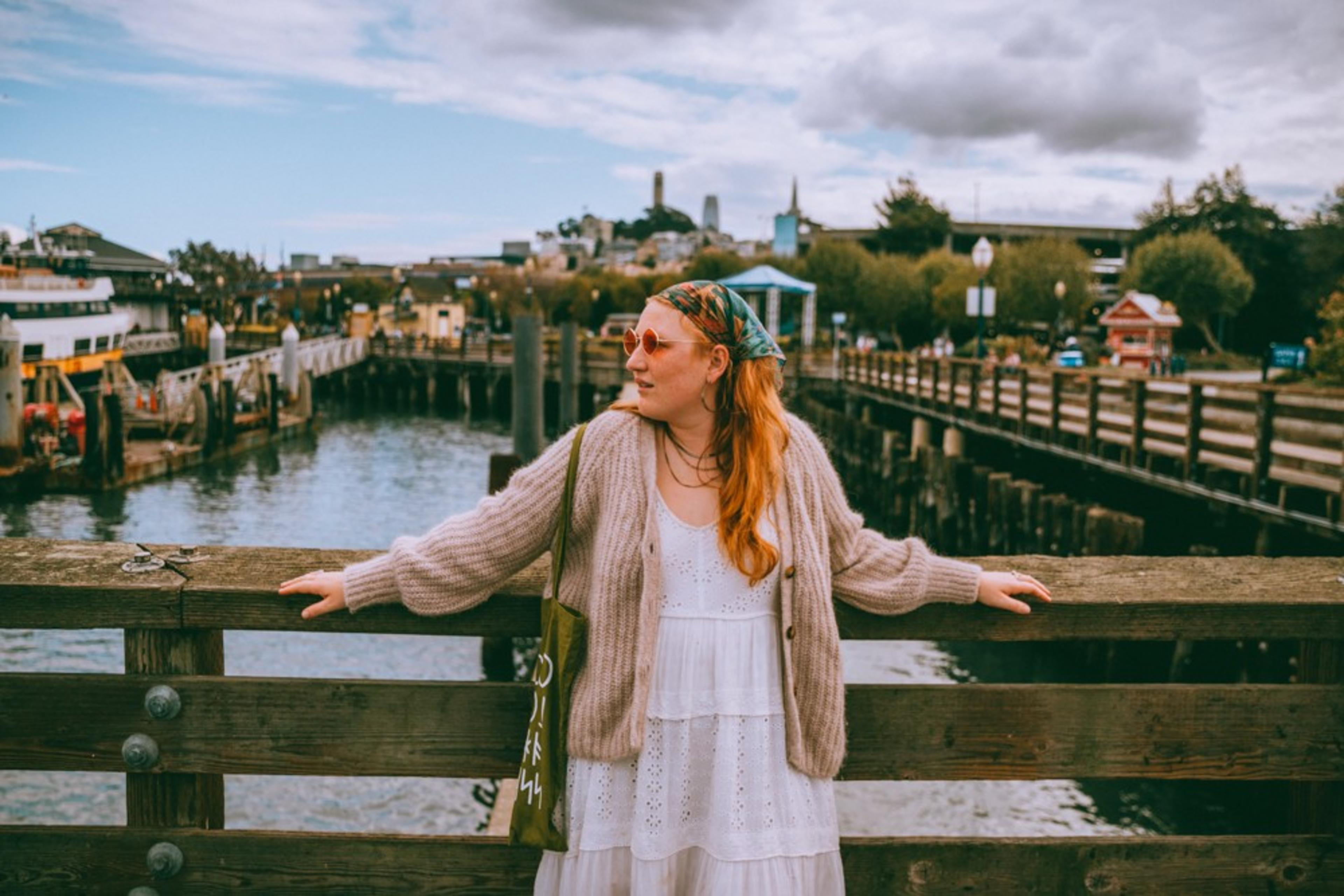 A woman in sunglasses leans on a wooden railing by the water at San Francisco's Fisherman's Wharf, with buildings and a cloudy sky in the background.