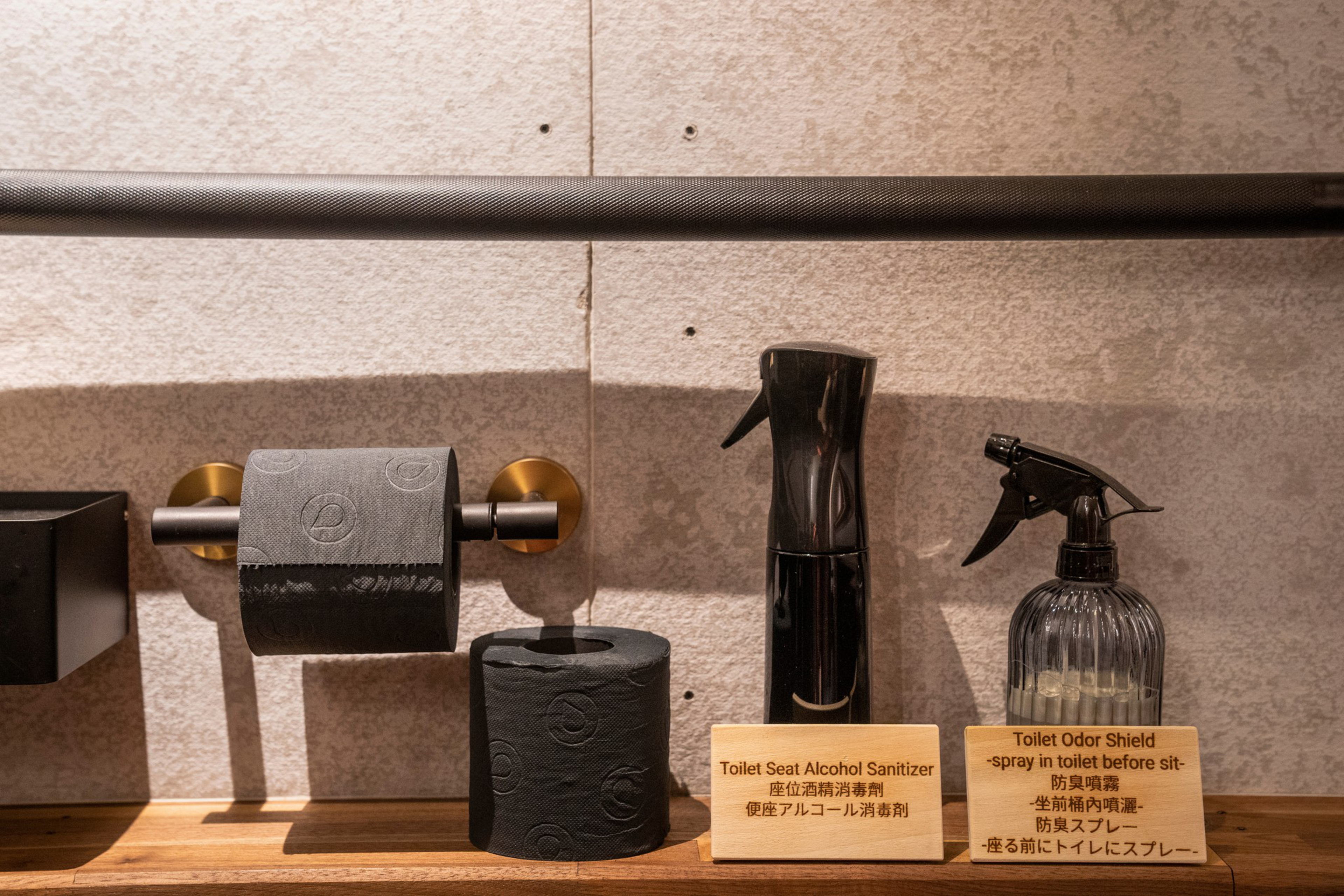 The image shows a bathroom setup with a black toilet paper holder, black toilet paper, and two labeled spray bottles for seat sanitizing and odor shielding.