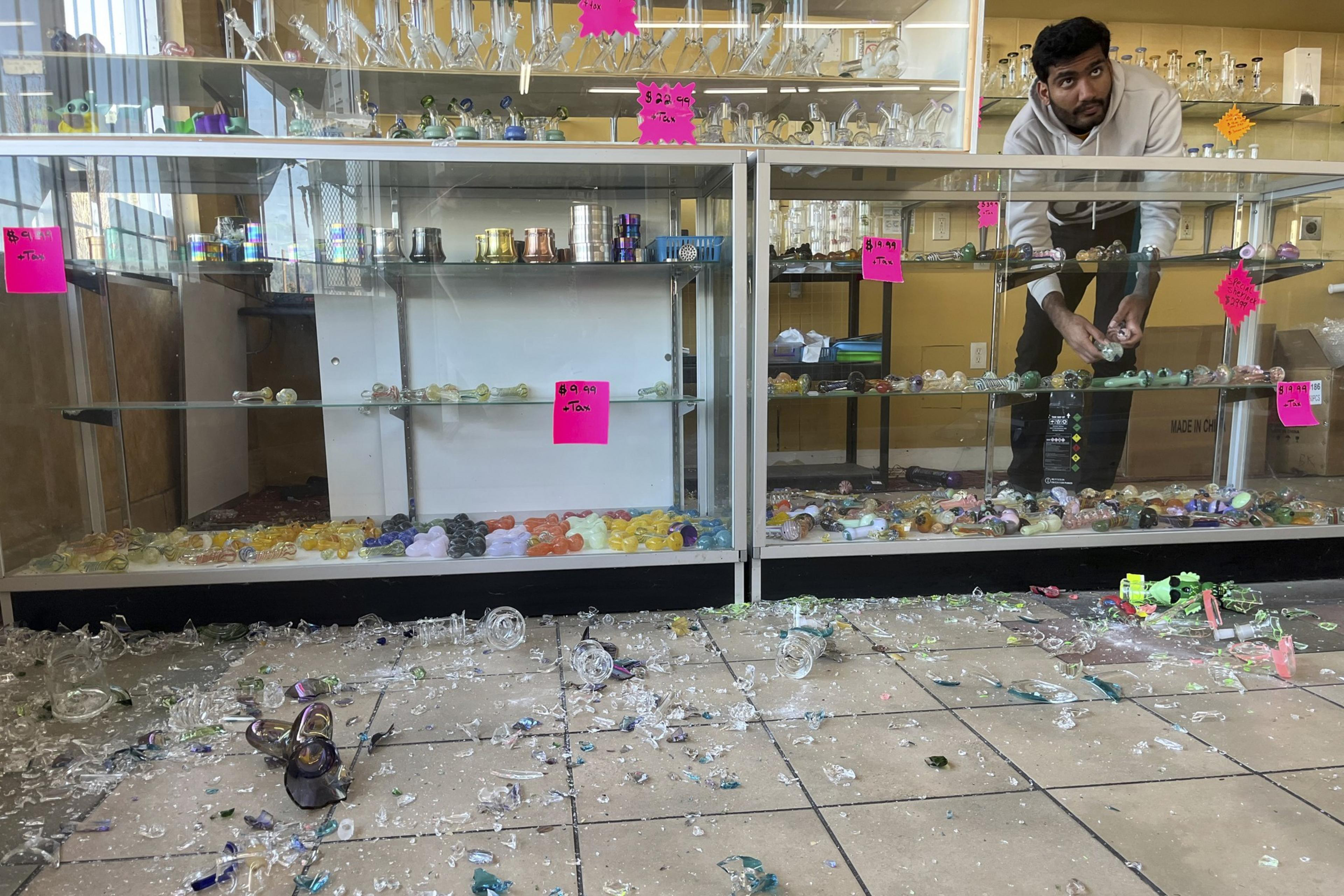 A man in a store gathers items from broken glass cases. Colorful glass pipes and pieces are scattered across the floor with bright signs on the cases.