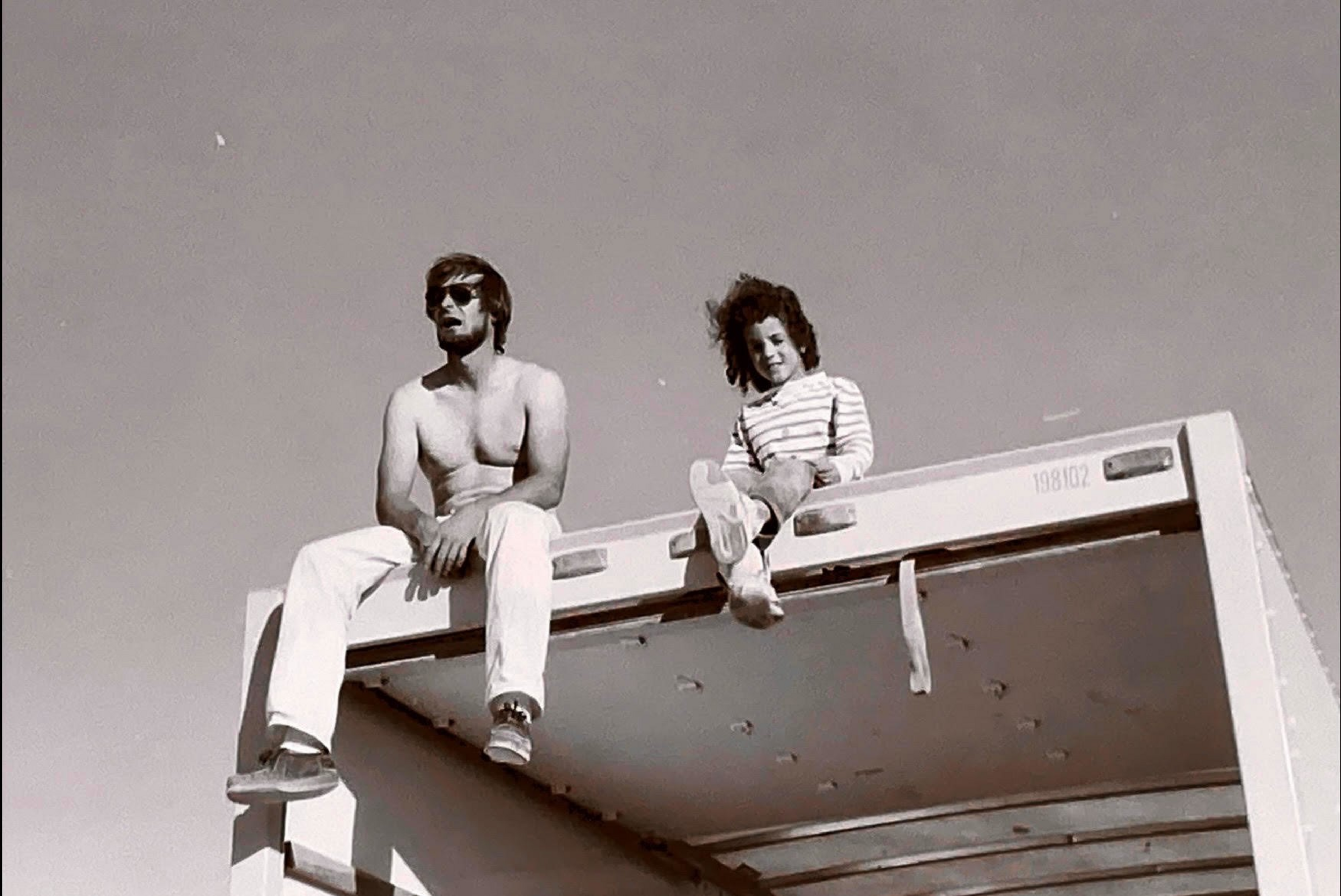 A shirtless man wearing sunglasses sits next to a girl in a striped long-sleeve shirt atop a high metal structure against a clear sky.