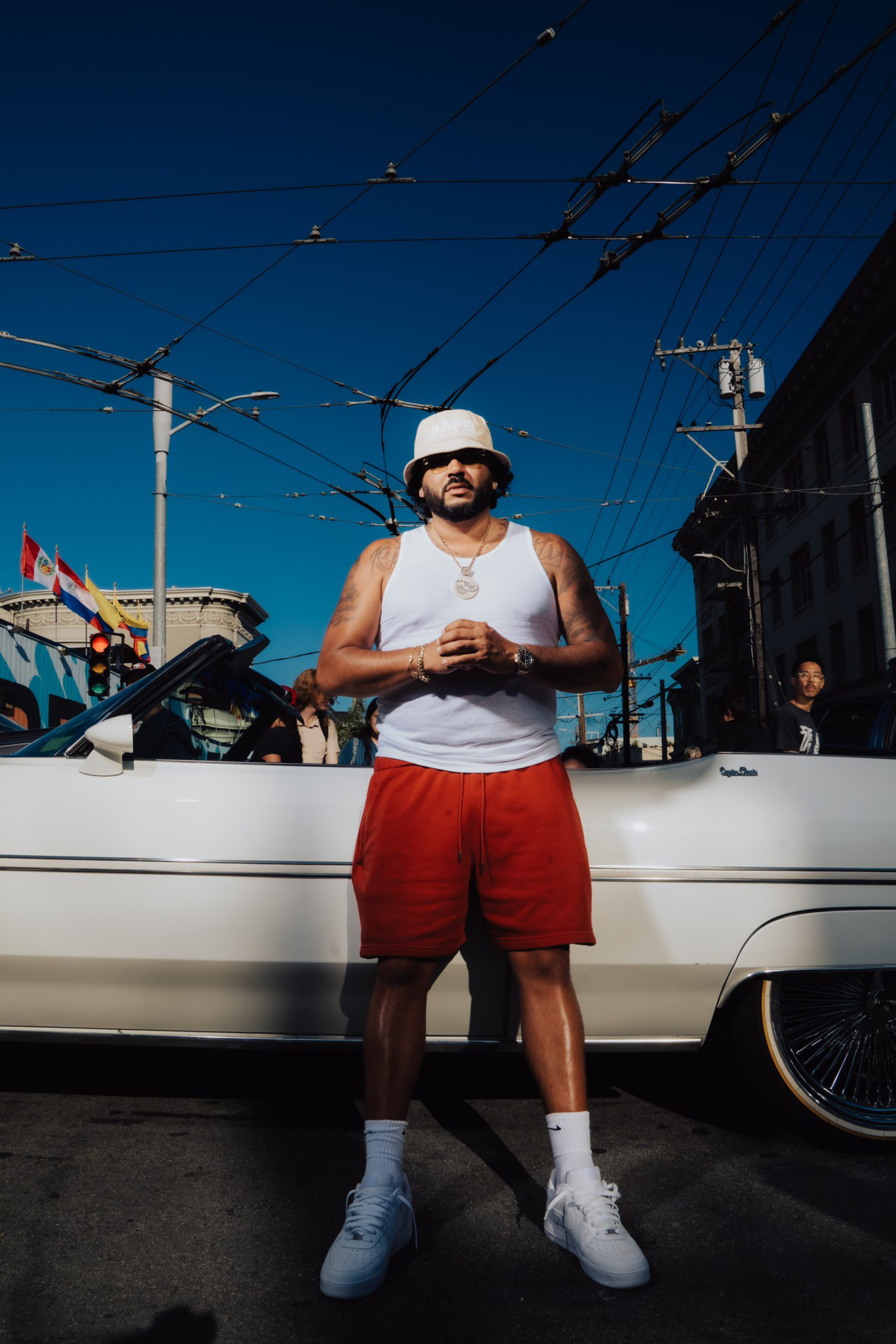 A man in a white tank top, red shorts, white bucket hat, and sneakers stands confidently in front of a white convertible car on a sunny street.