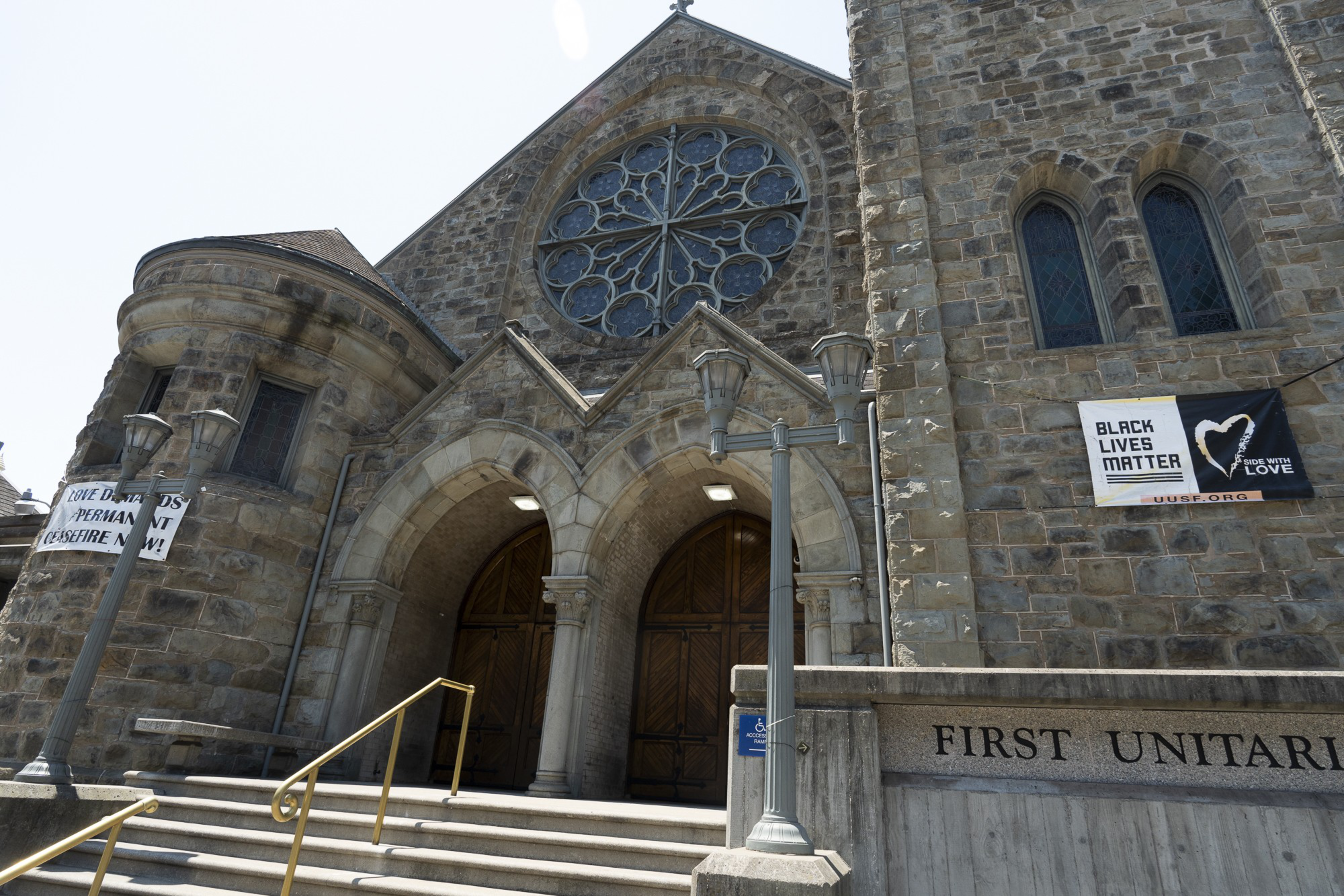 A stone church entrance features wooden doors and a large circular stained glass window. Banners express support for Black Lives Matter and a ceasefire.