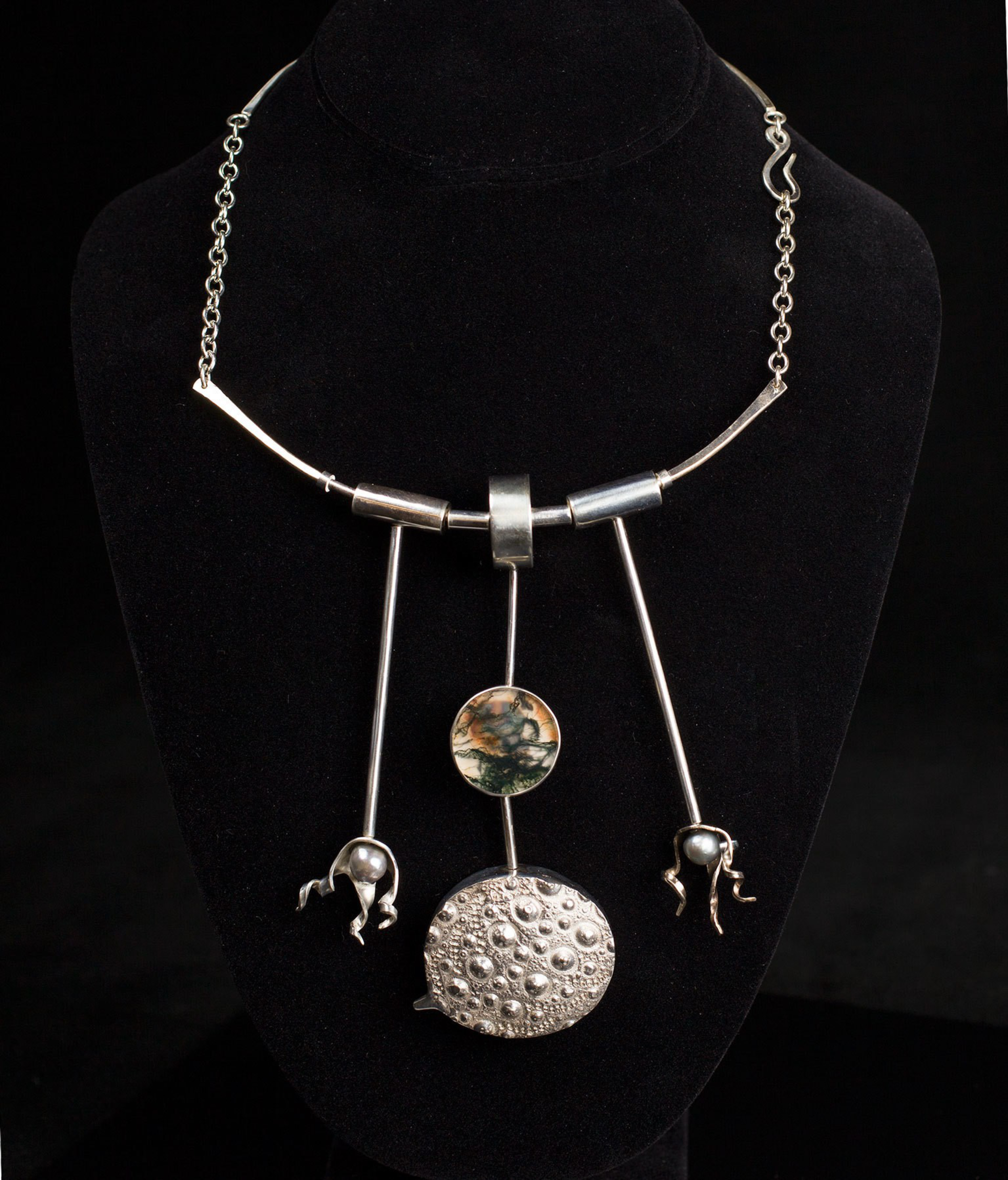 A silver necklace with three vertical rods, one holding a round stone with green and brown patterns, and two holding round textured silver pendants with small spheres.