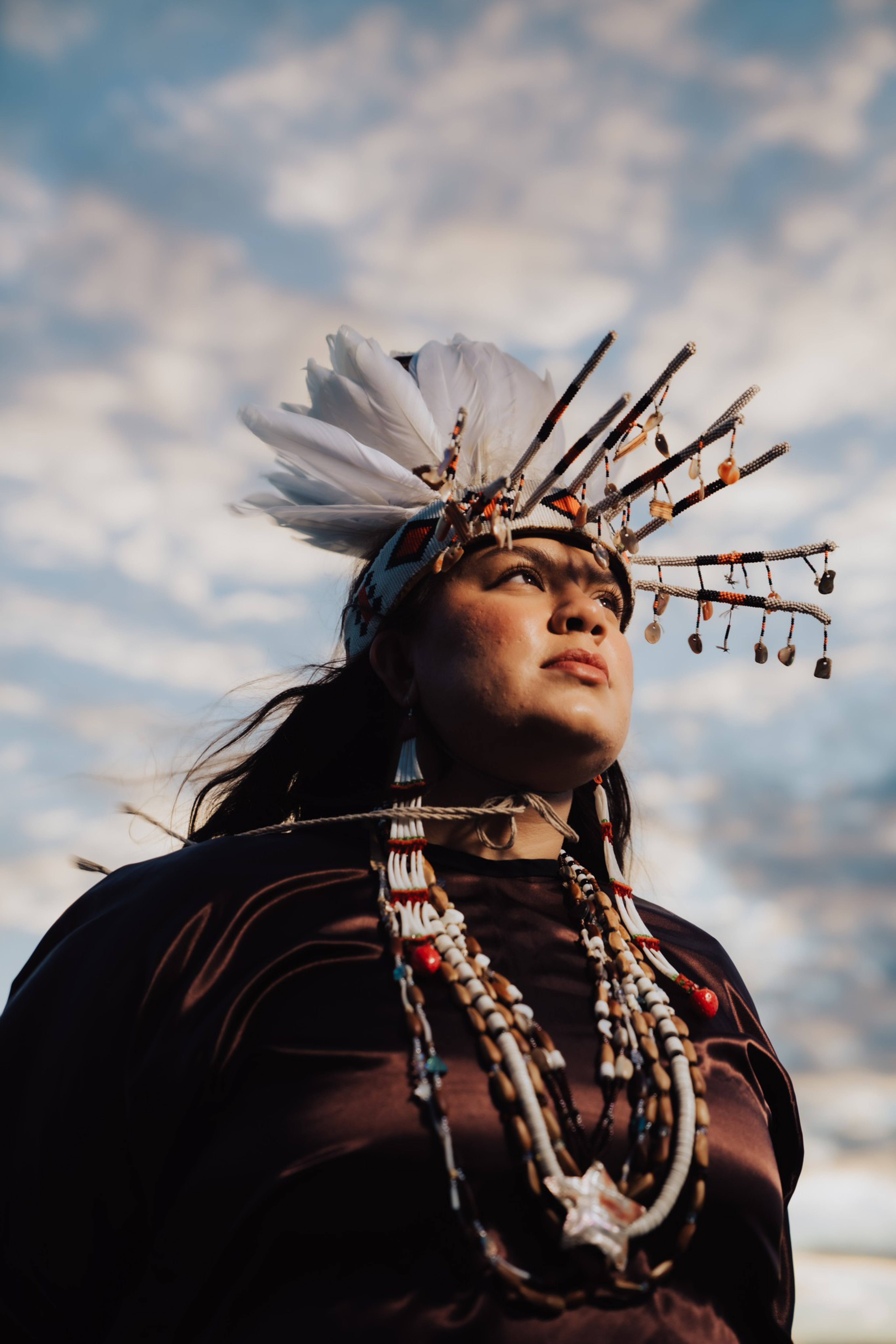 A person wearing a traditional headdress with white feathers and beads looks thoughtfully into the distance against a cloudy sky.