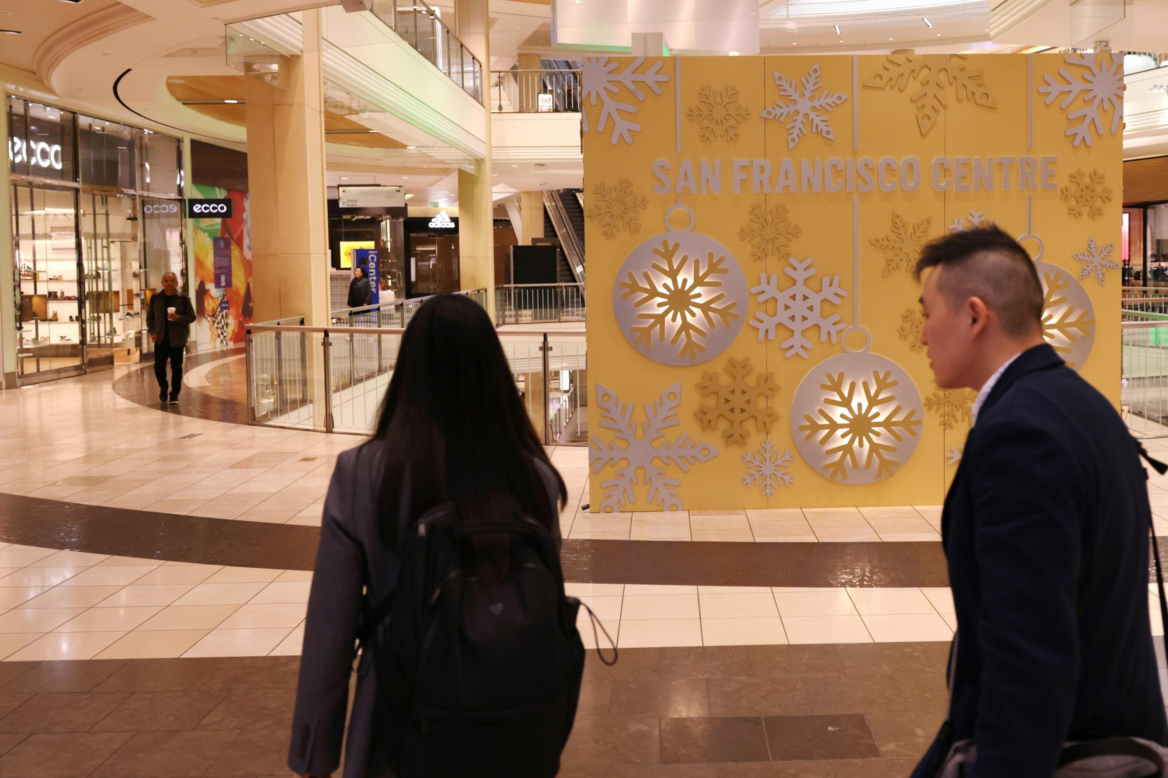 A shopping center with people walking by a large yellow sign displaying "SAN FRANCISCO CENTRE" adorned with snowflakes.