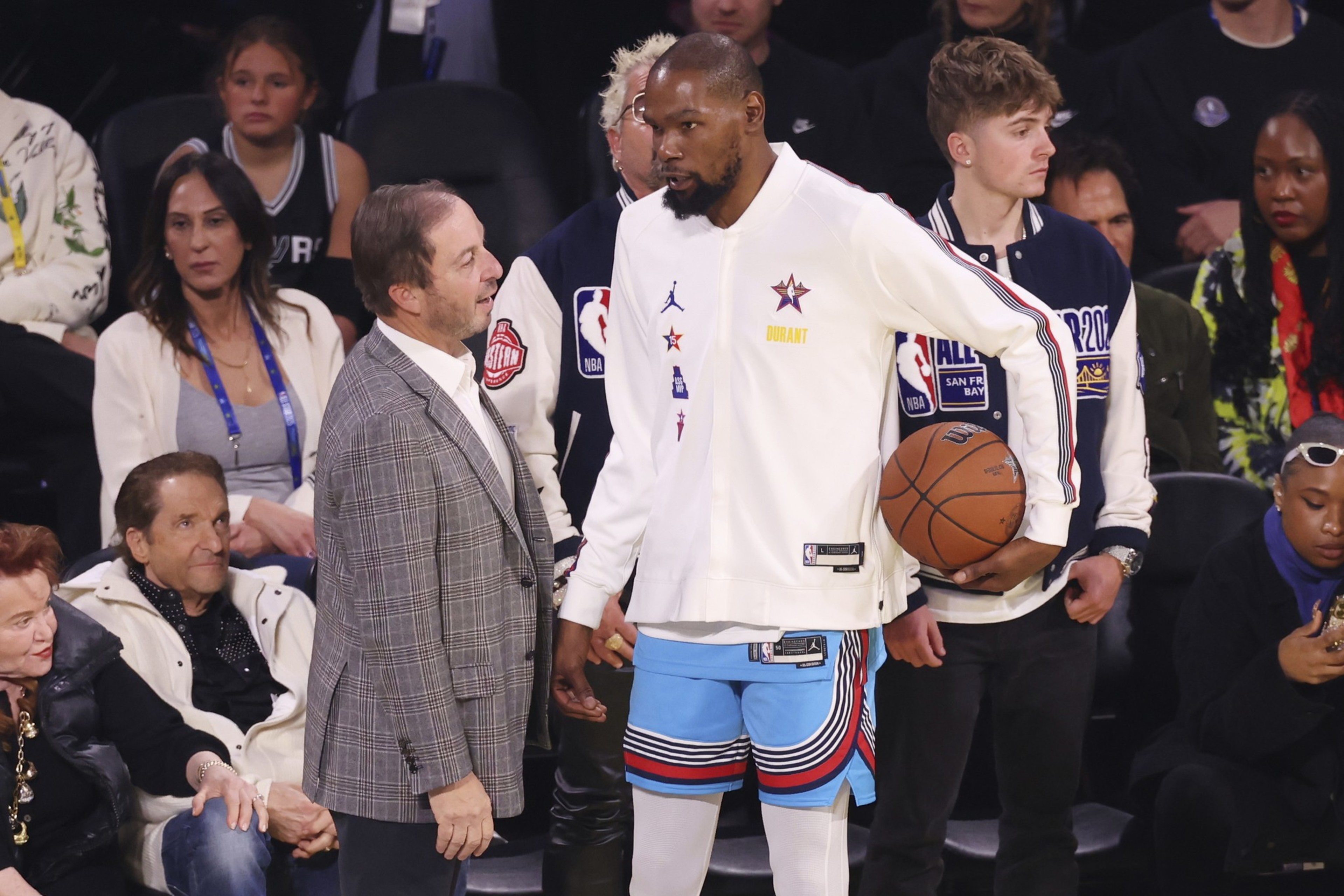 A basketball player in a white jersey with &quot;Durant&quot; on it holds a ball while talking to a man in a plaid suit. People are seated and standing around them.