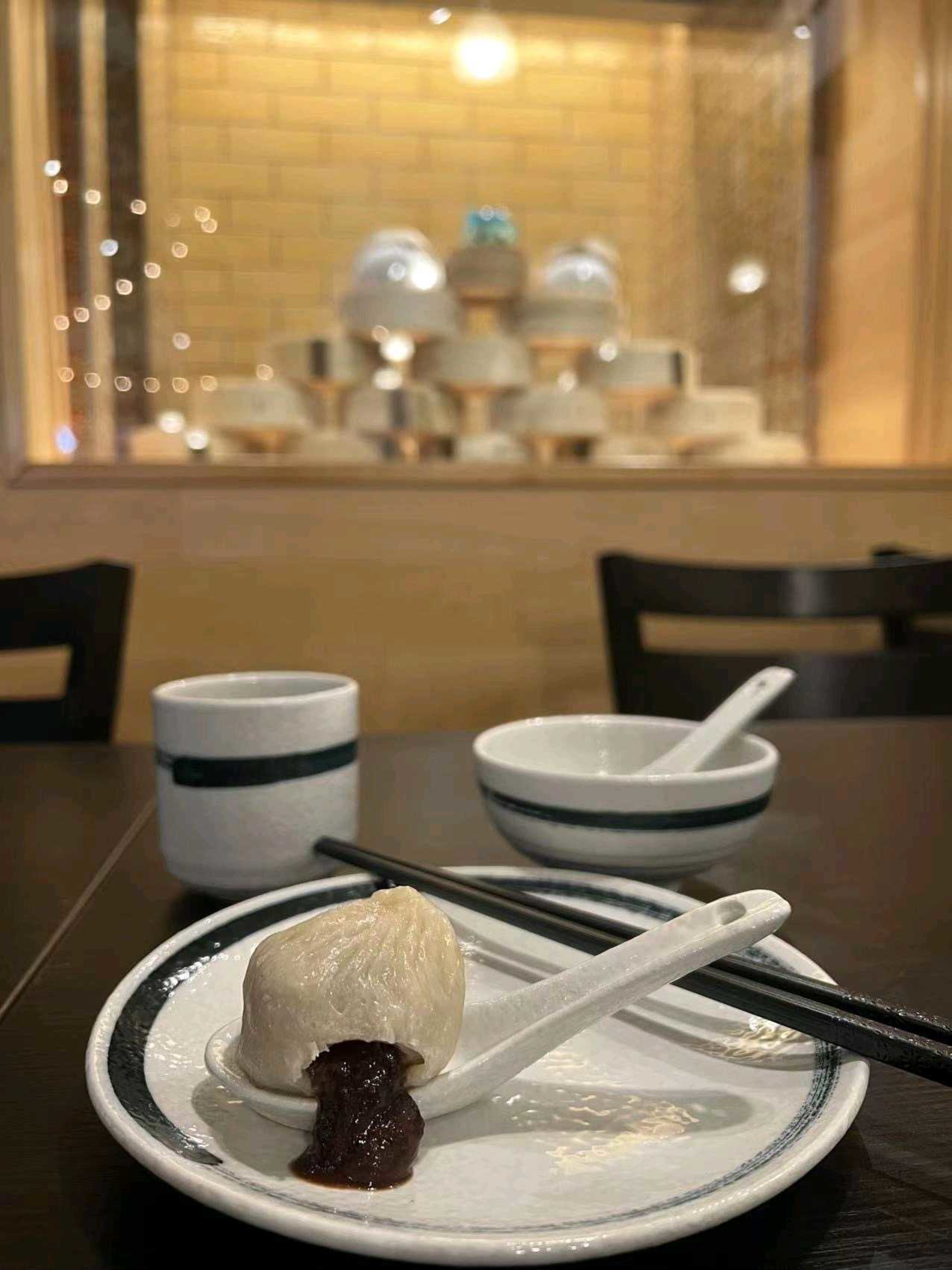 A steamed bun with chocolate filling oozing out rests on a ceramic spoon atop a white plate with black trim, accompanied by chopsticks, a cup, and a bowl.