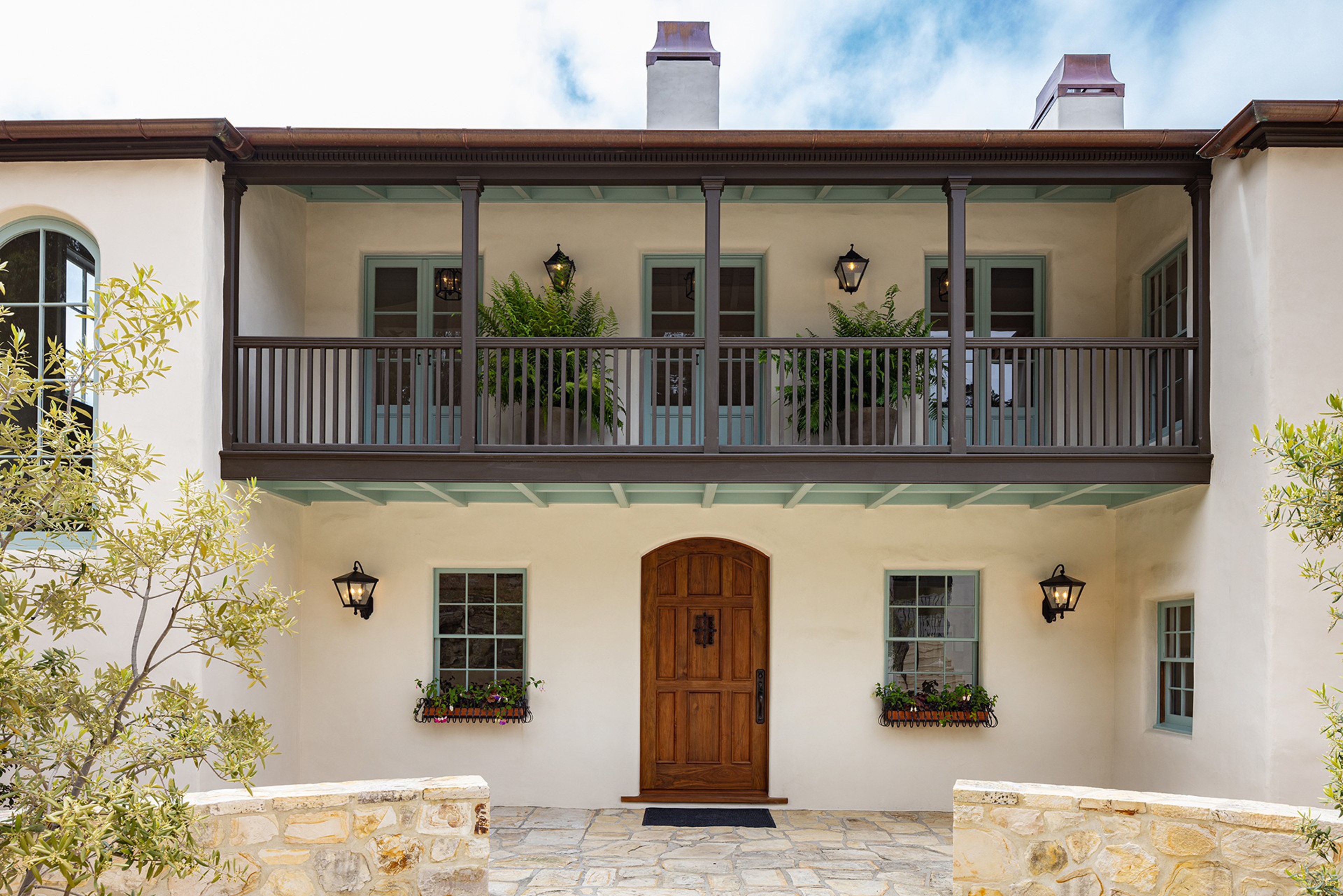A two-story house with a wooden front door, stone pathway, balcony with two potted plants, lantern-style wall lights, and flower boxes under two windows.