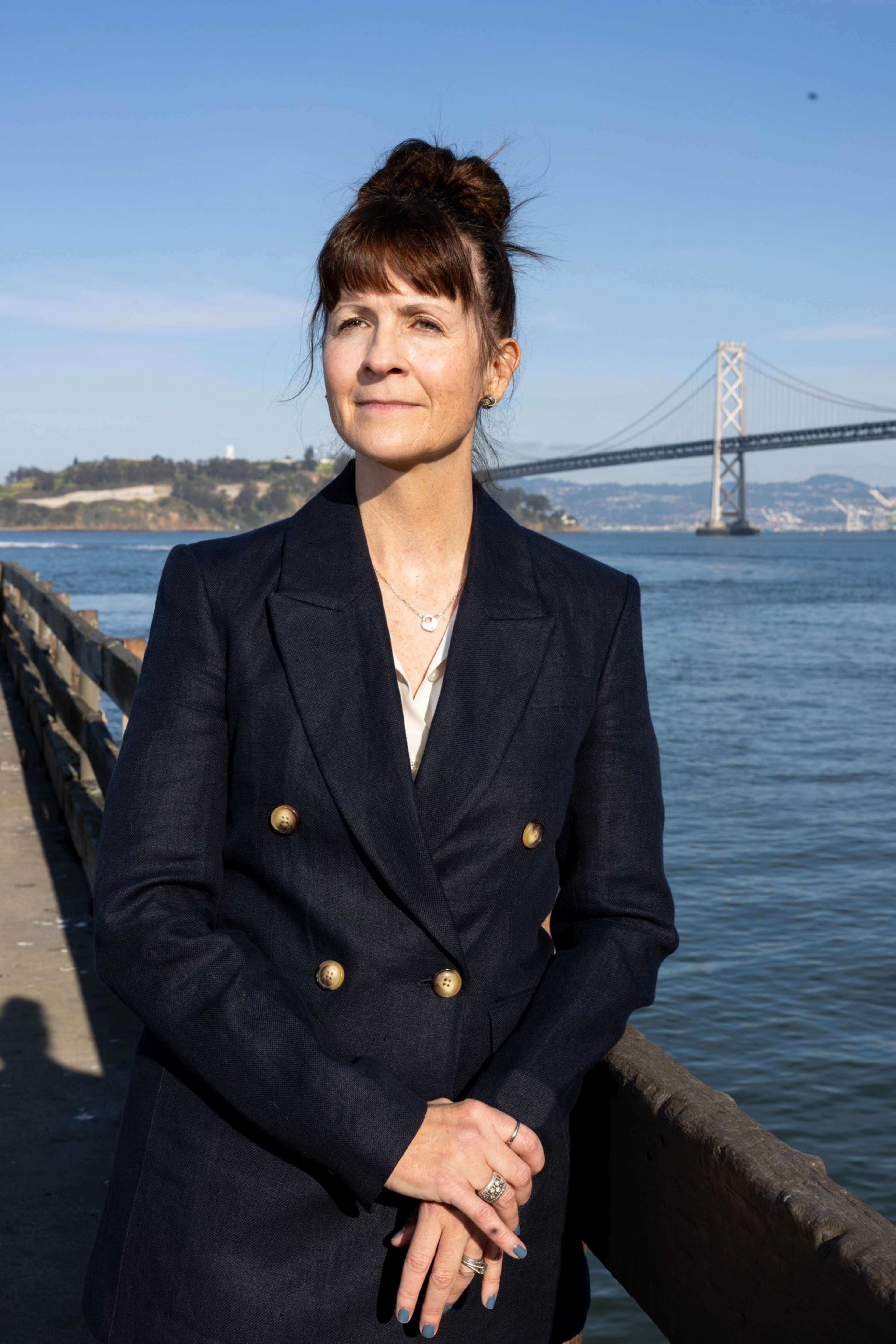 A woman in a dark double-breasted blazer stands by a waterfront railing with a large suspension bridge and clear sky in the background.