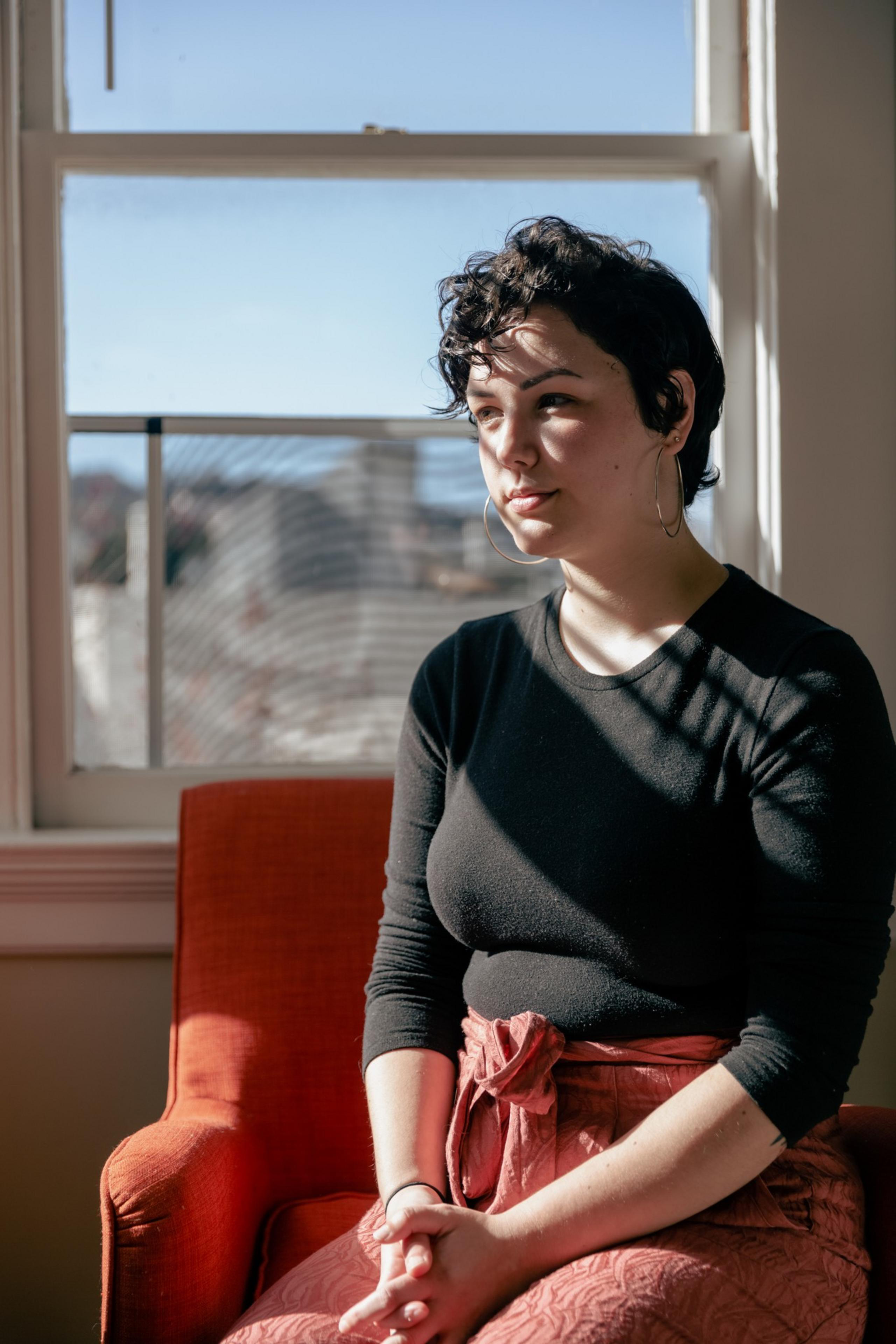 Sasha Perigo sits by a window, sunlight casting shadows, in thoughtful pose, wearing a black top and hooped earrings.