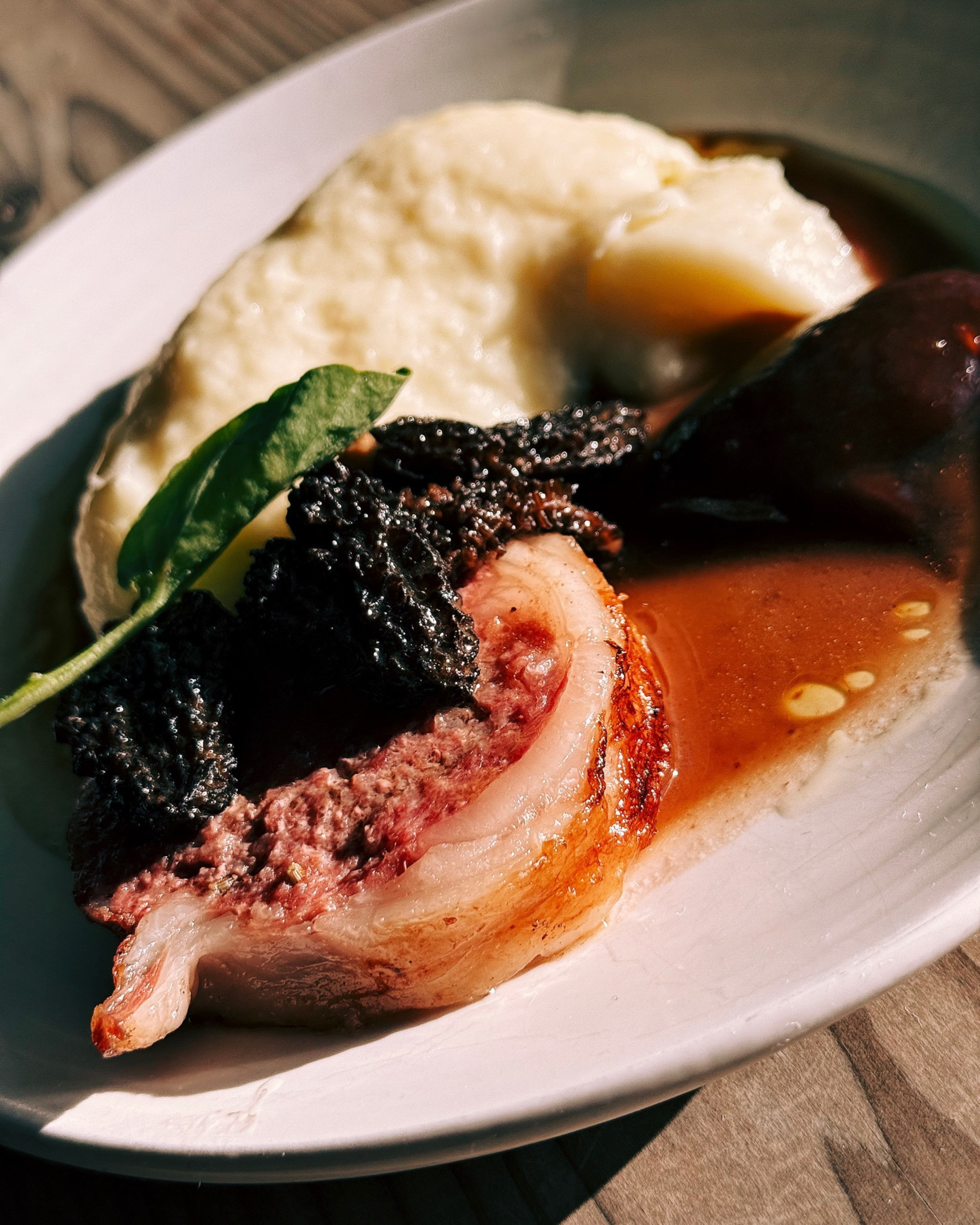 A dish featuring seared meat roulade topped with mushrooms, accompanied by creamy mashed potatoes and a rich brown sauce, garnished with a green leaf.