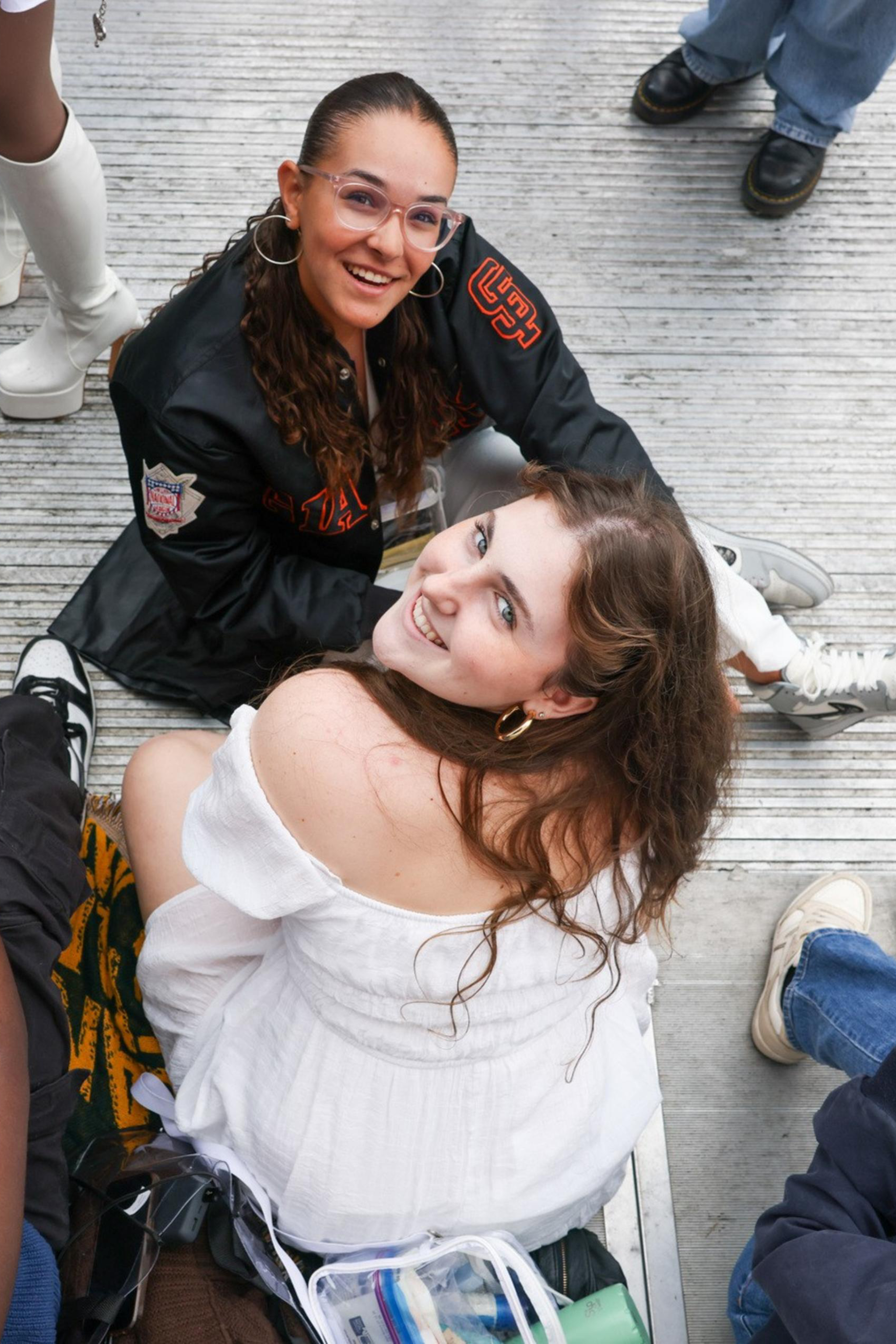 The image shows a group of people sitting on the ground, engaged in various activities. Two women are looking up and smiling towards the camera, while others are using their phones.