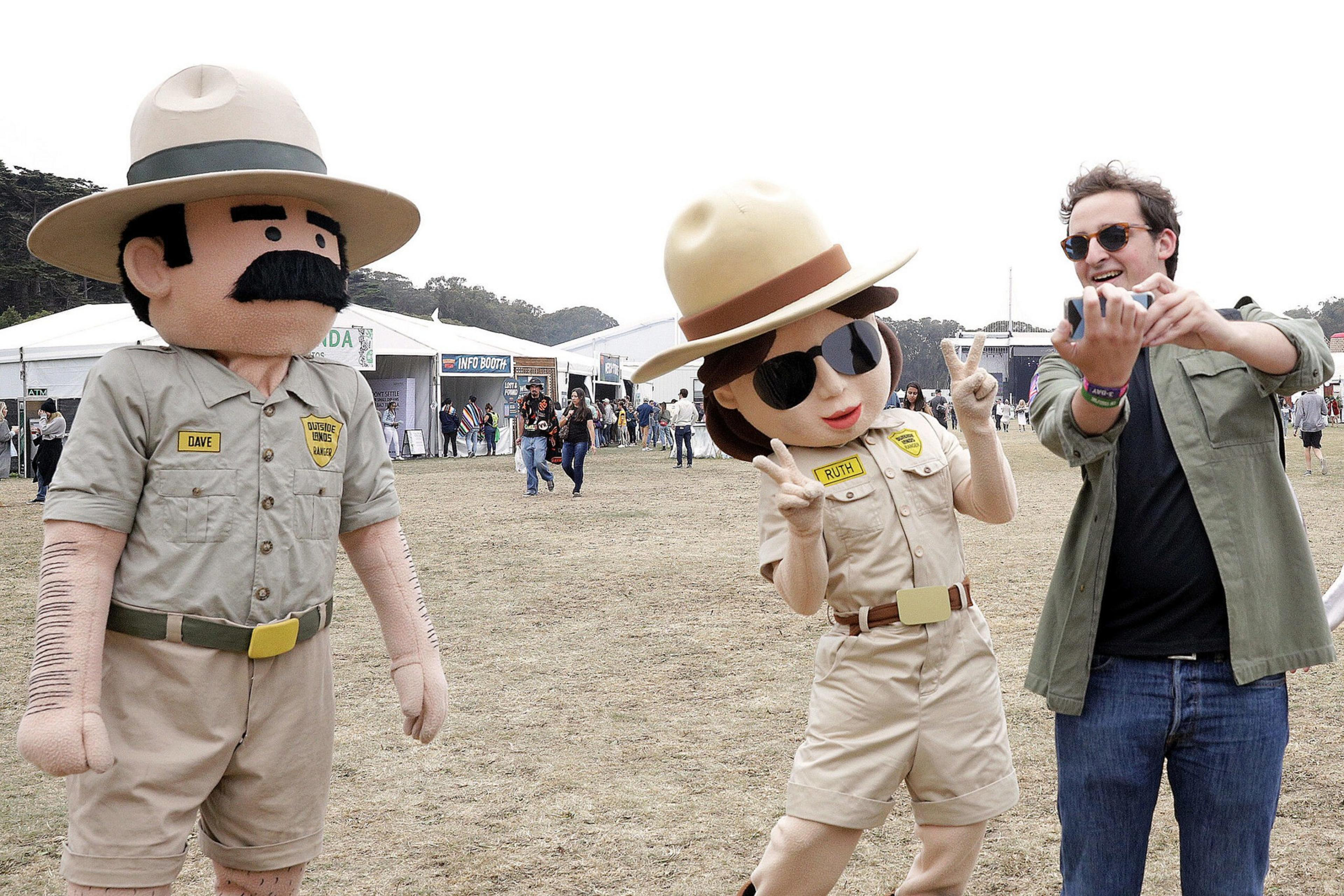 Two people dressed in oversized park ranger costumes pose with a man wearing sunglasses, who is taking a selfie on a grassy open field near tents.