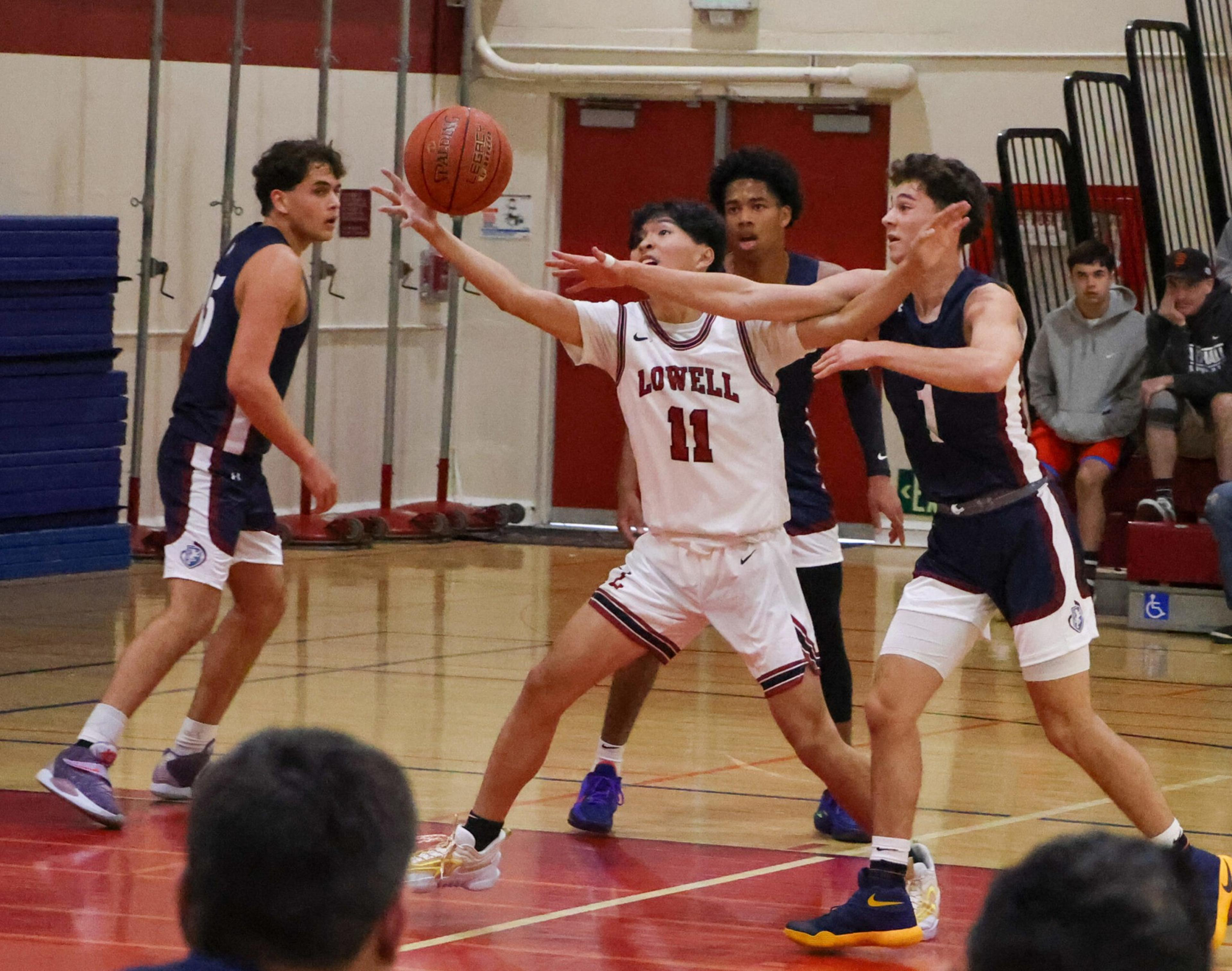 A basketball player in a white Lowell jersey stretches to catch the ball while three players in dark jerseys try to block him in a gym.
