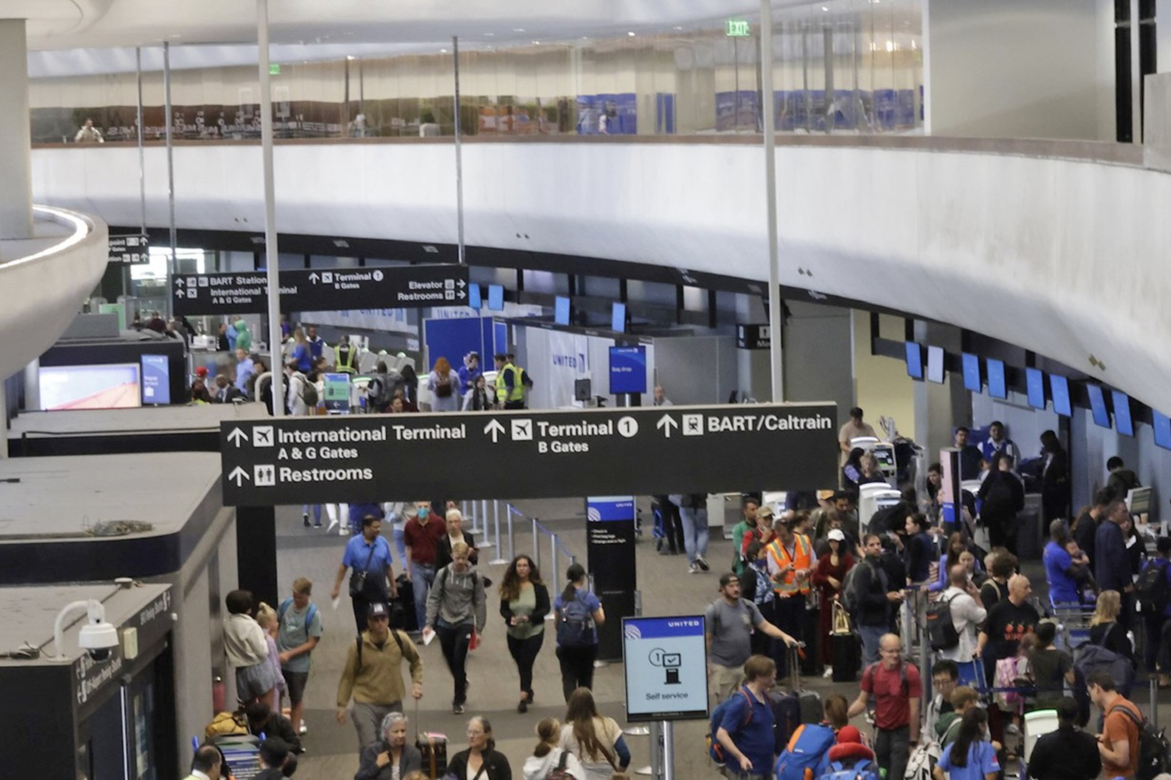 The image shows a busy airport terminal with many people walking around, checking in, and waiting in line. There are overhead signs directing travelers to various gates, terminals, and restrooms.