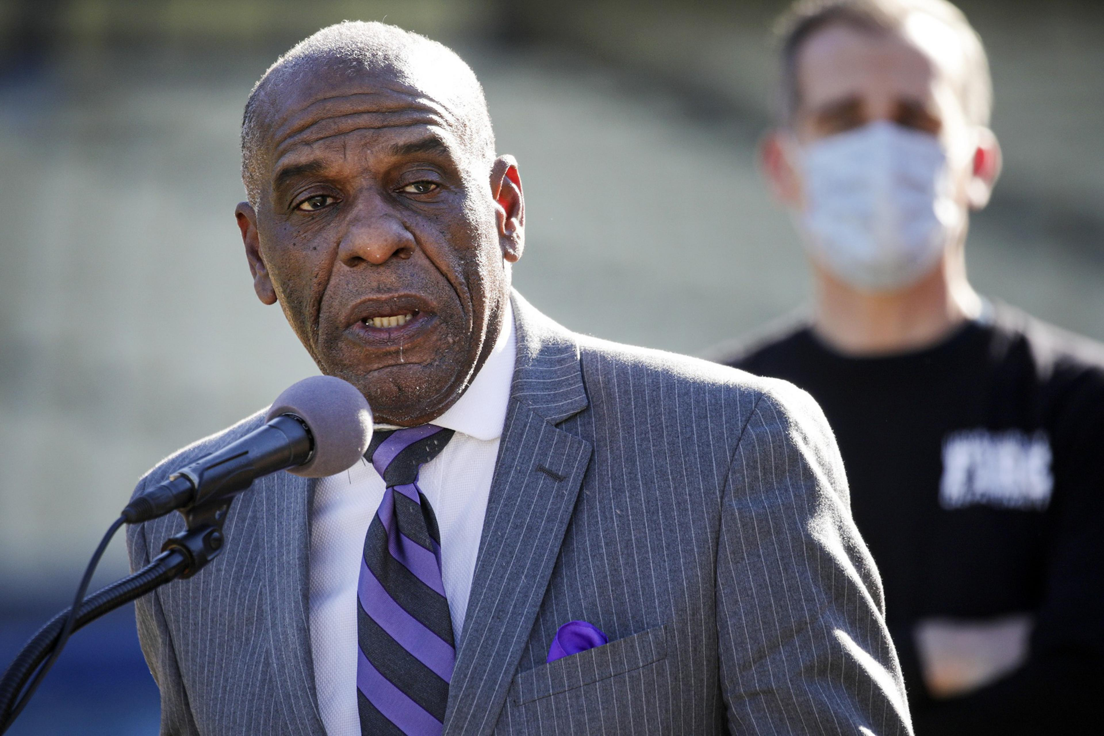 A man in a suit speaks at a microphone; a masked person stands behind him.