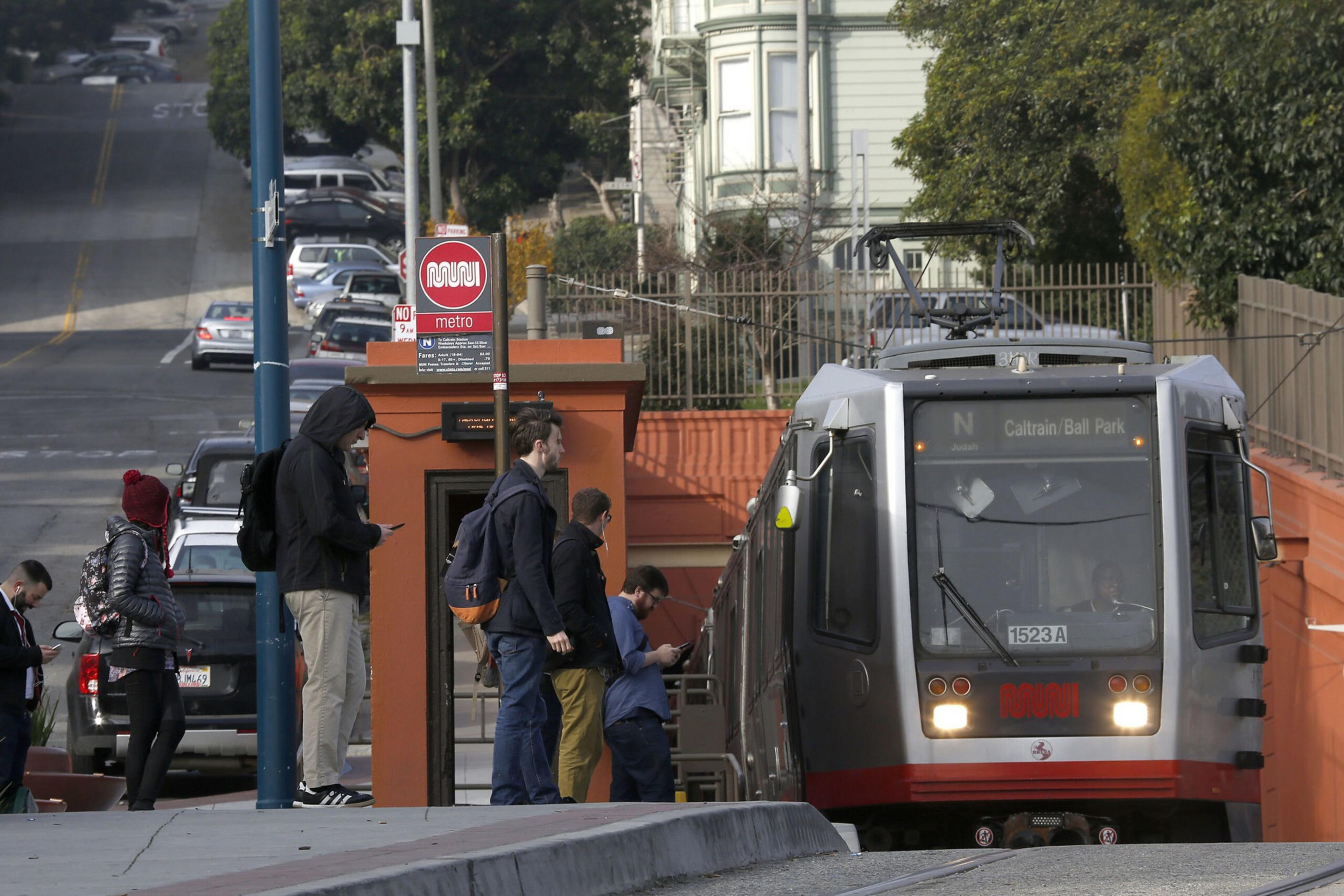 People wait at a metro station platform as a gray and red train labeled "Caltrain/Ball Park" approaches.