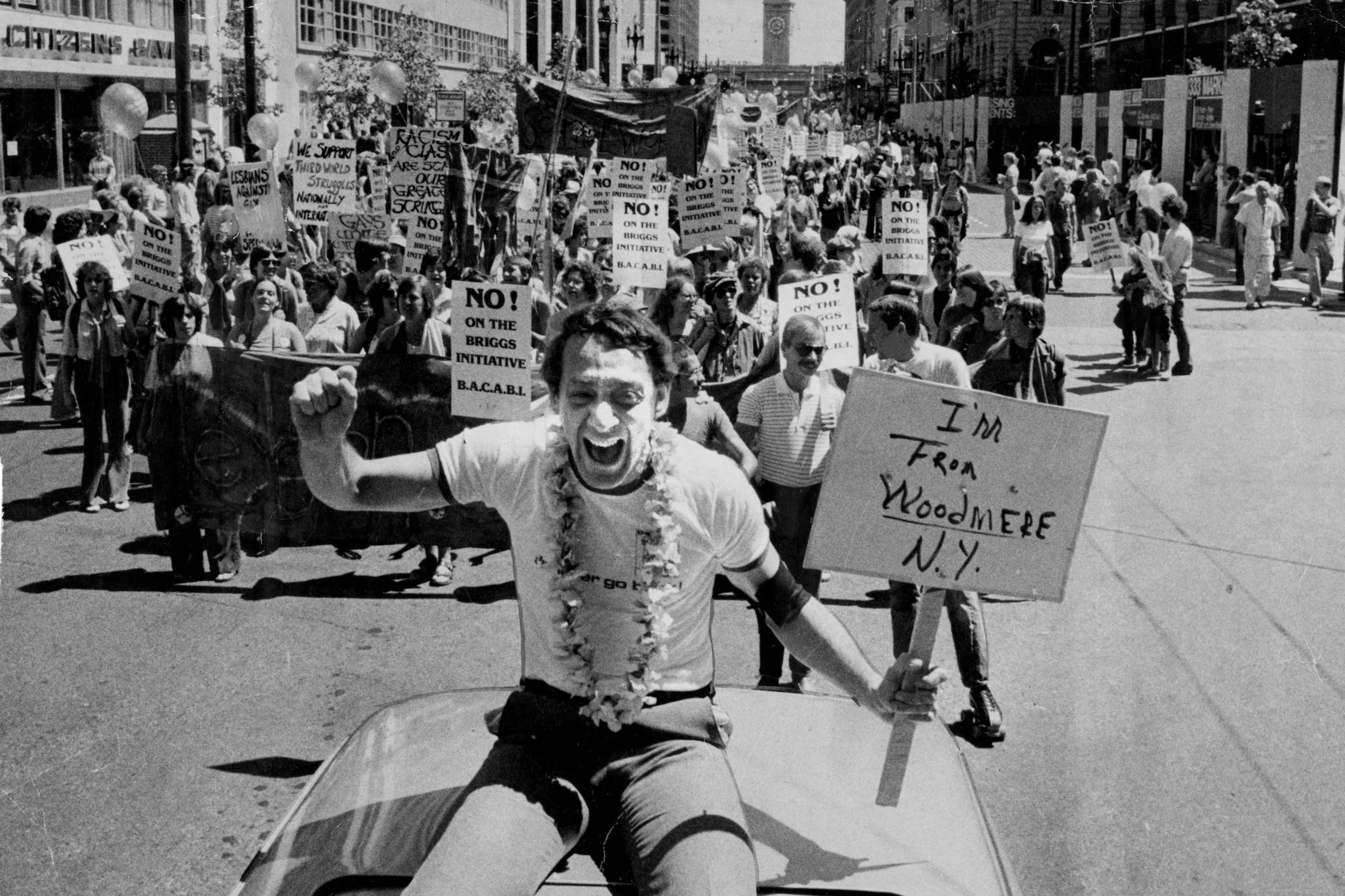 A man wearing a lei holds a sign saying "I'm from Woodmere N.Y." while leading a large protest with many holding signs opposing the Briggs Initiative.