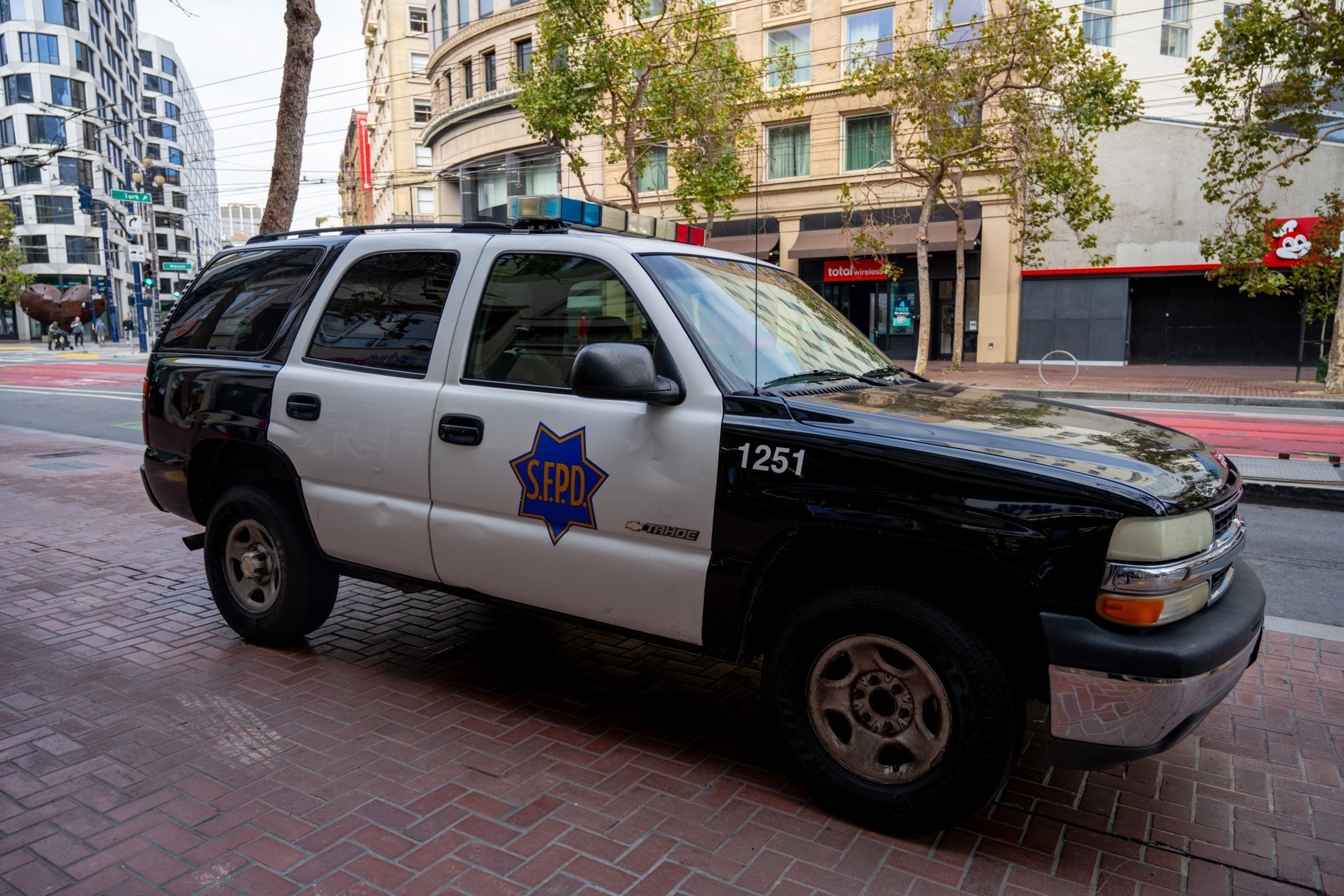 A parked black and white S.F.P.D. Tahoe police SUV with the number 1251 on the front door sits on a brick sidewalk in an urban area.