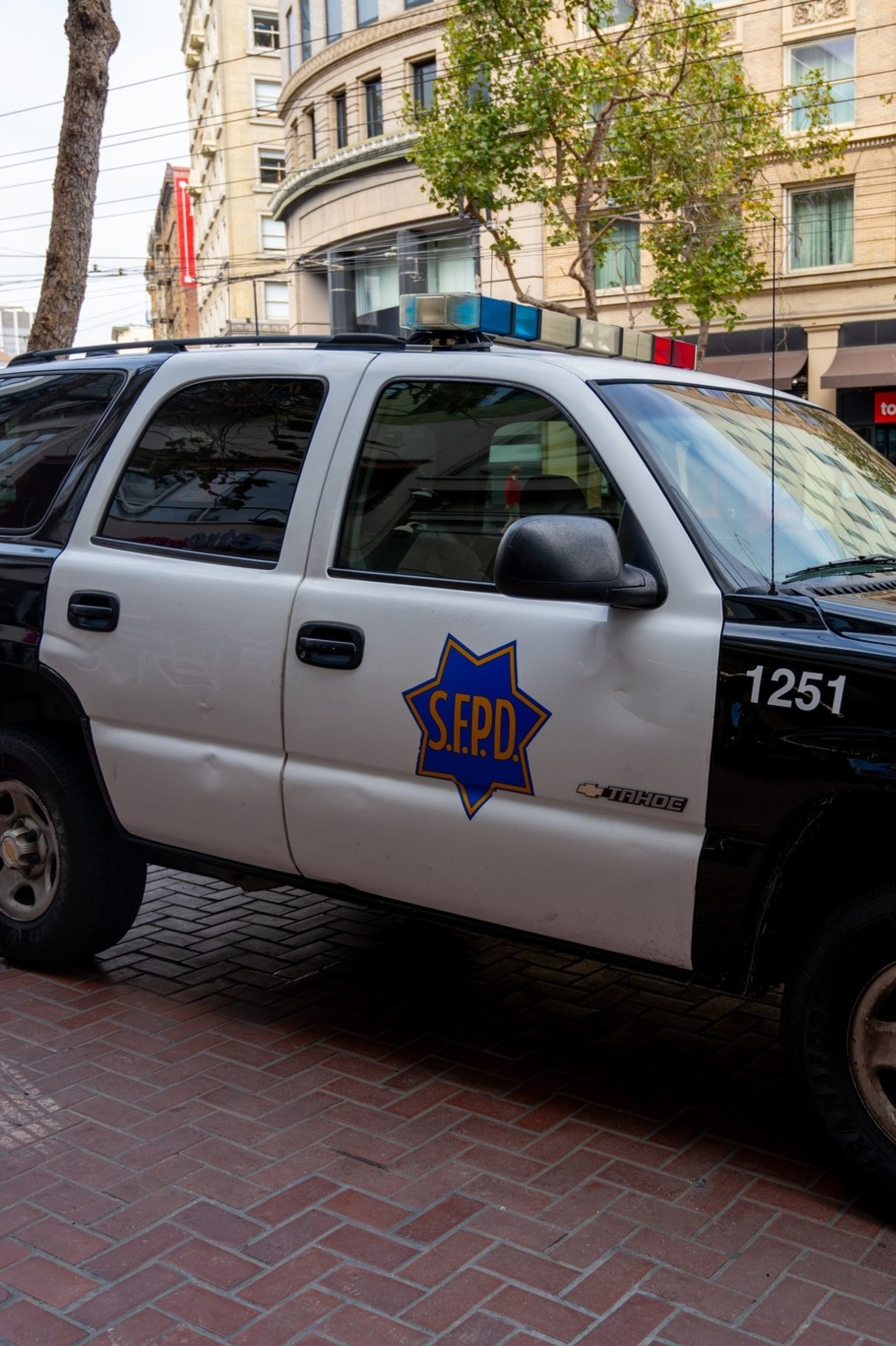 A parked black and white S.F.P.D. Tahoe police SUV with the number 1251 on the front door sits on a brick sidewalk in an urban area.