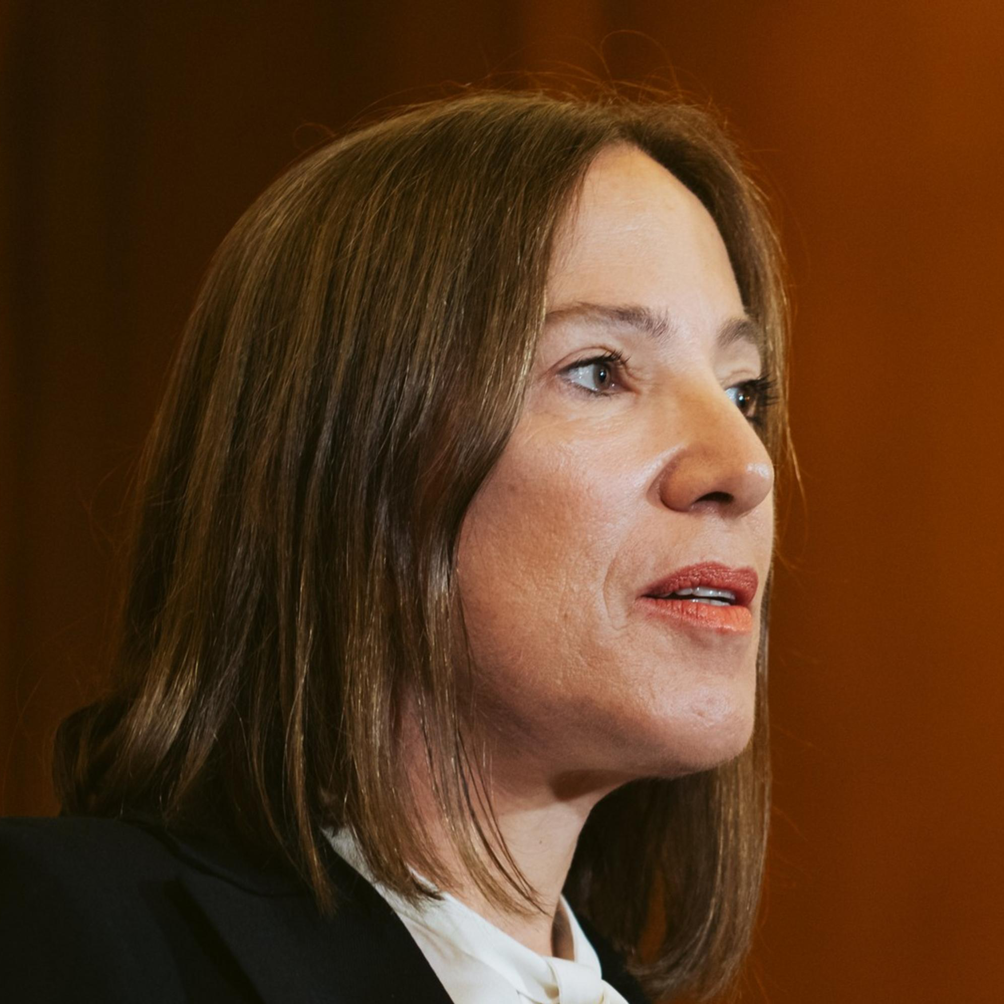 A woman with straight brown hair is speaking into a black microphone. She is wearing a white blouse and a black blazer. The background is brown with a slight blur.