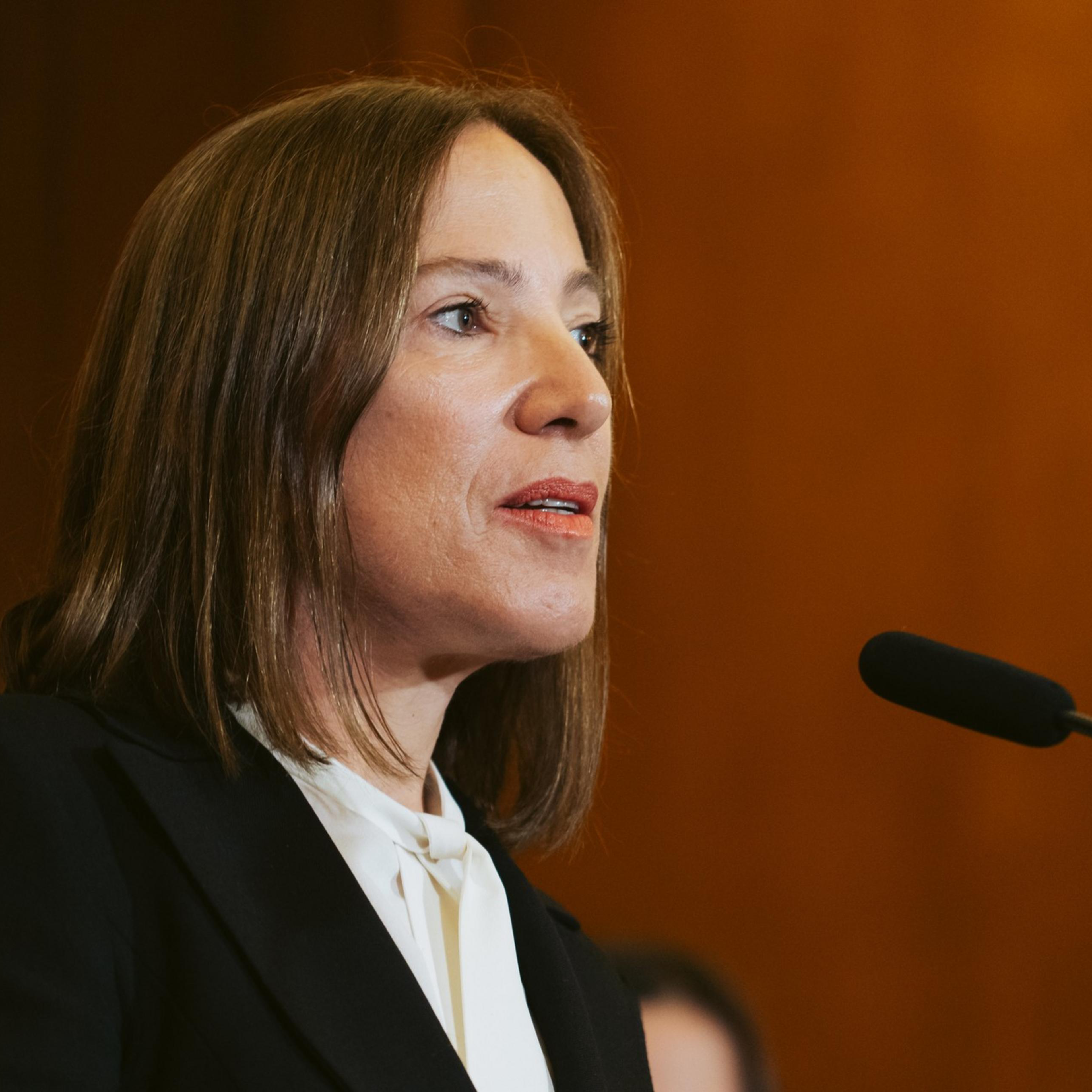 A woman with straight brown hair is speaking into a black microphone. She is wearing a white blouse and a black blazer. The background is brown with a slight blur.