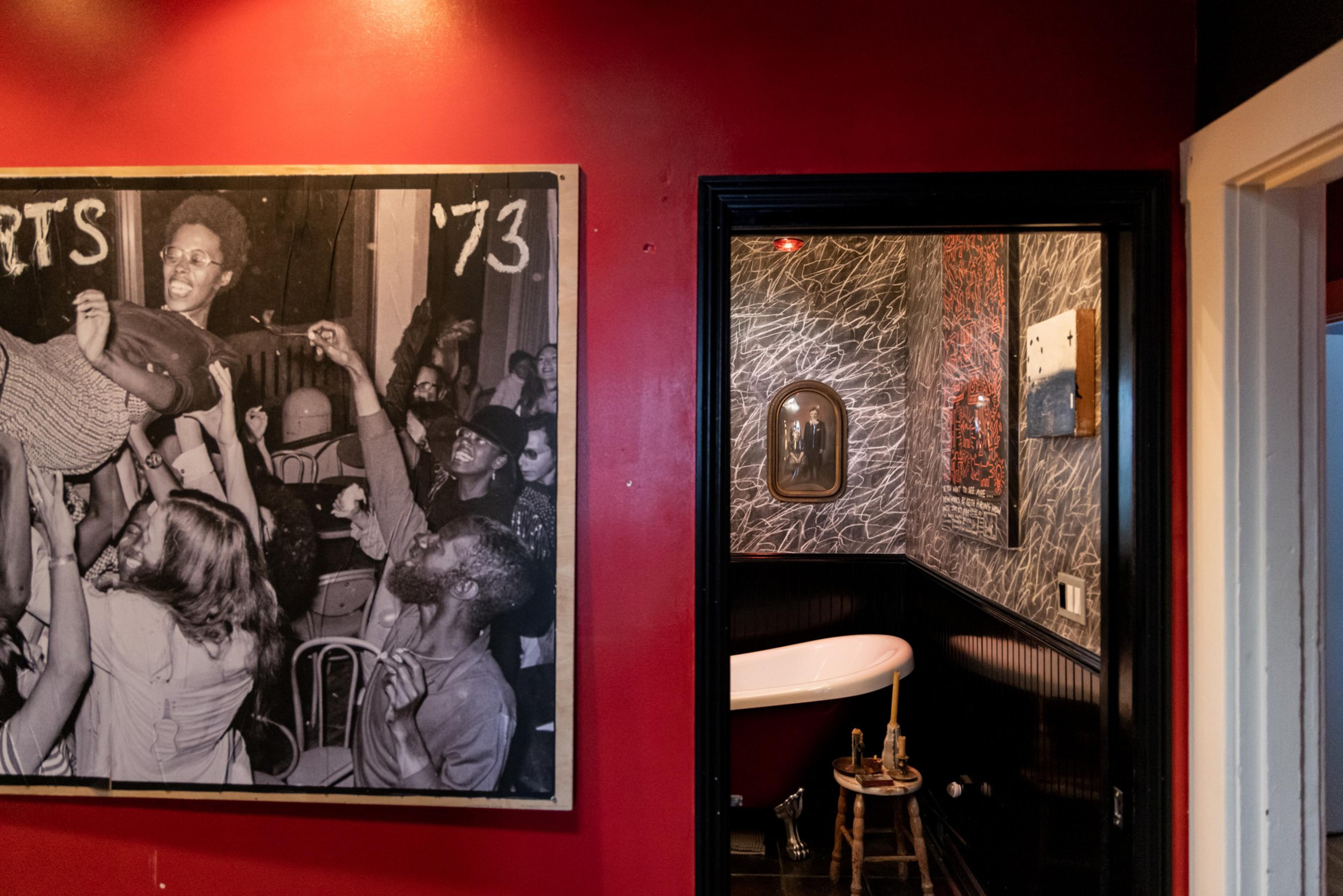 The image shows a red-walled room with a black-and-white photo of people celebrating in 1973. A doorway leads to a small, stylish bathroom with a clawfoot bathtub.