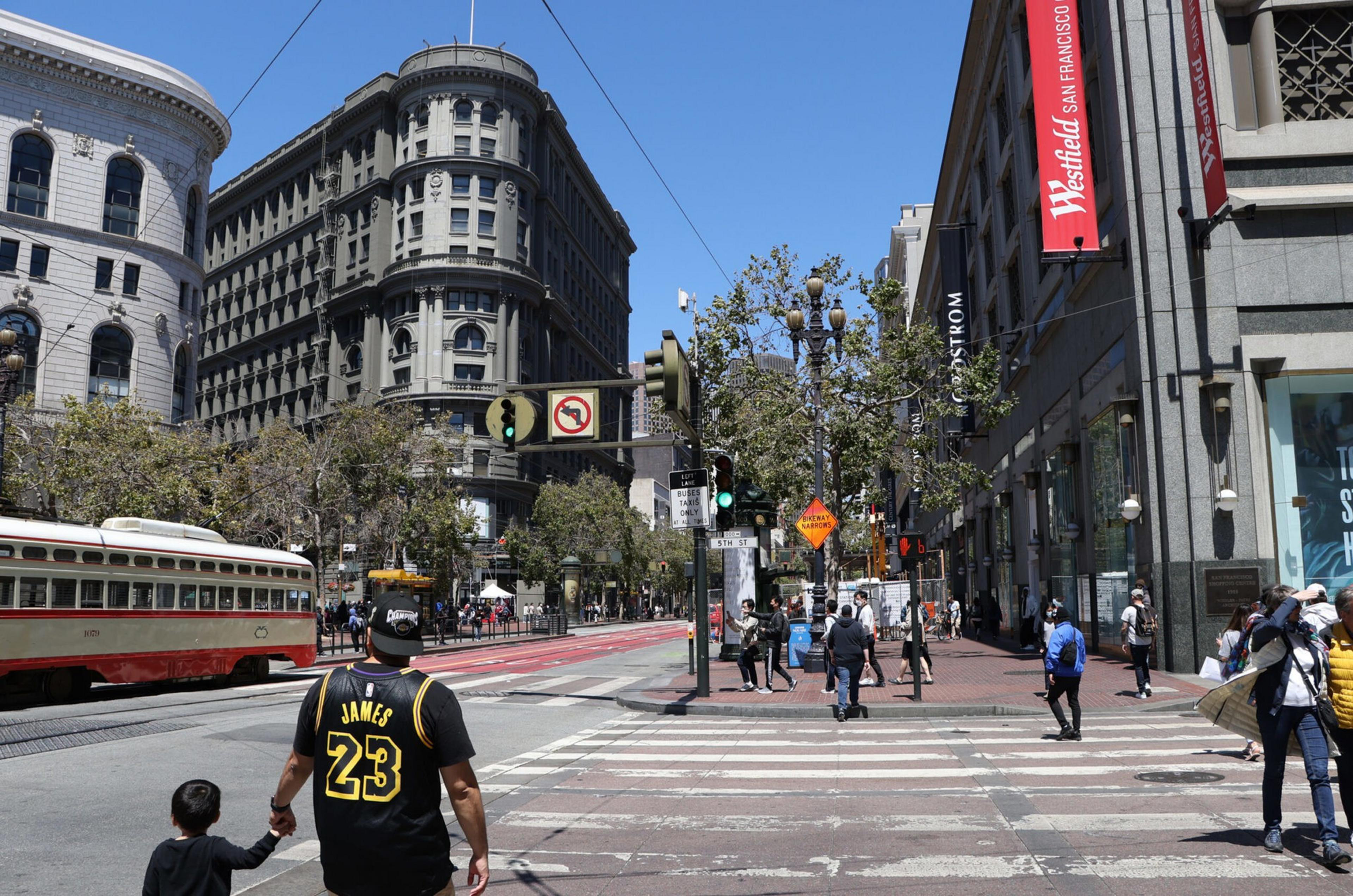 A man wearing a black and yellow "James 23" jersey holds a child's hand while crossing a street near a Westfield San Francisco mall and a red streetcar.