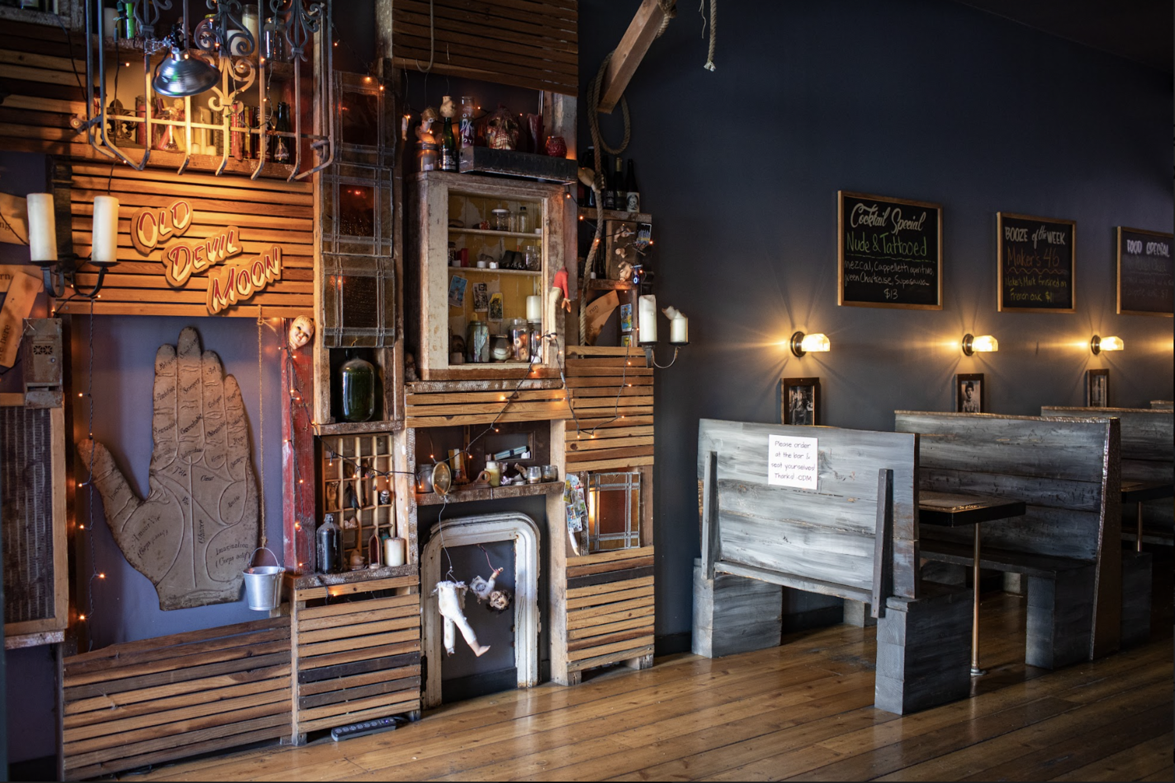 A cozy bar interior with wooden booths, warm lighting, shelves filled with trinkets, a large wooden hand art piece, and a neon sign reading "Old Devil Moon."