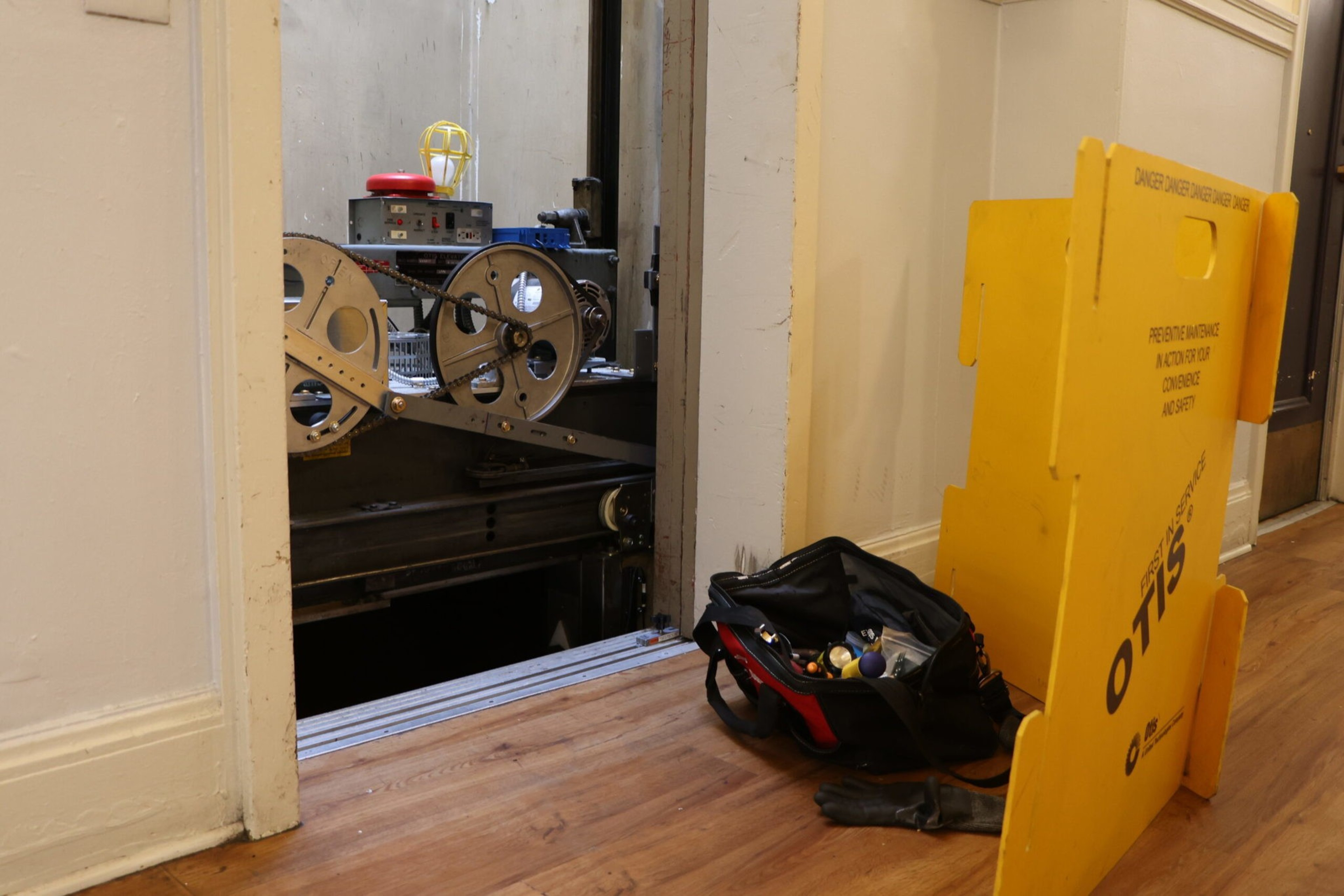 An open elevator shaft reveals its mechanical gears, with a toolbag, glove, and a yellow safety barrier labeled "OTIS" placed nearby on the wooden floor.