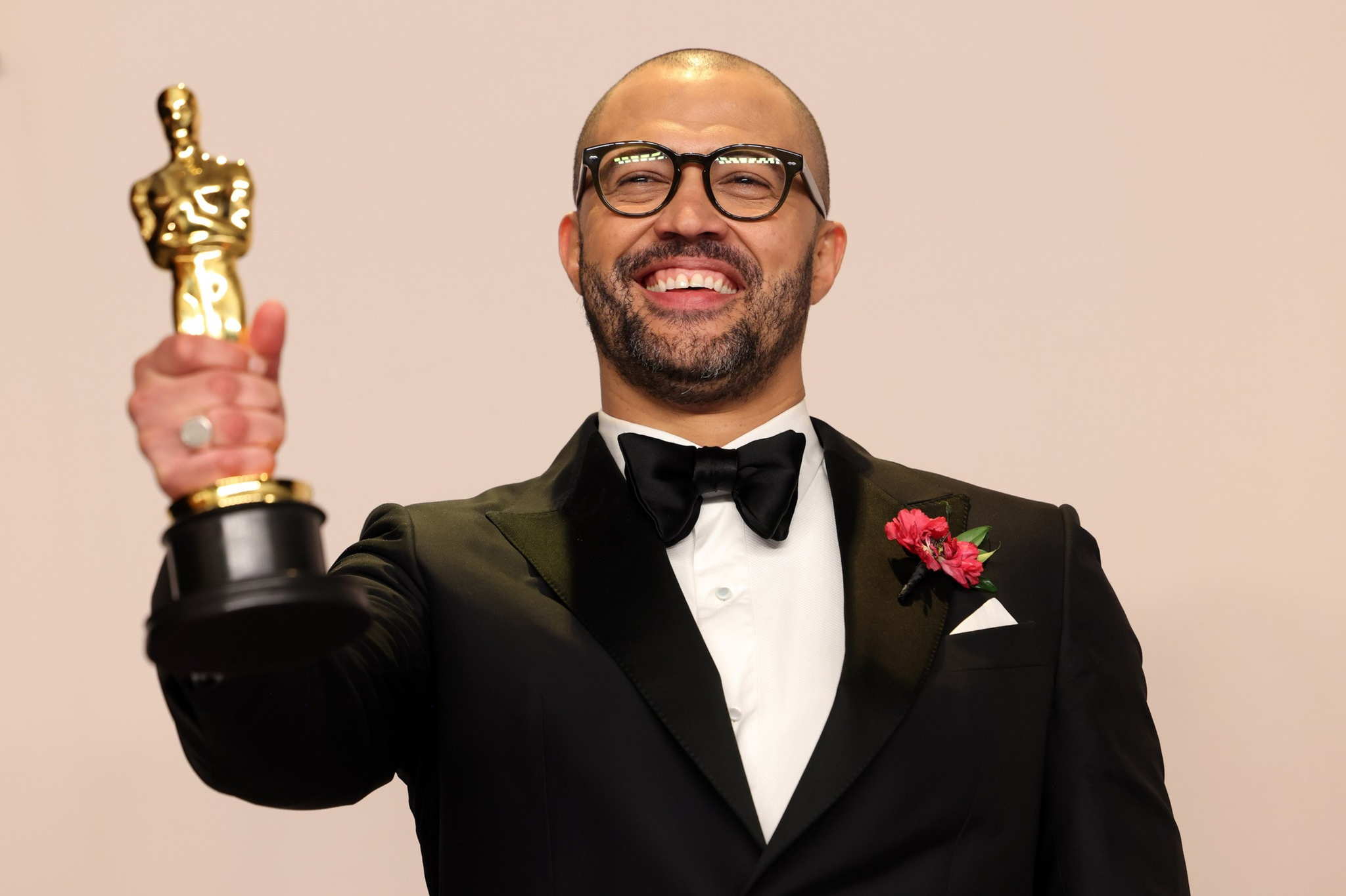 A smiling man in a tuxedo holds up an Oscar trophy. He wears glasses and has a boutonniÃ¨re with red flowers on his lapel against a plain background.