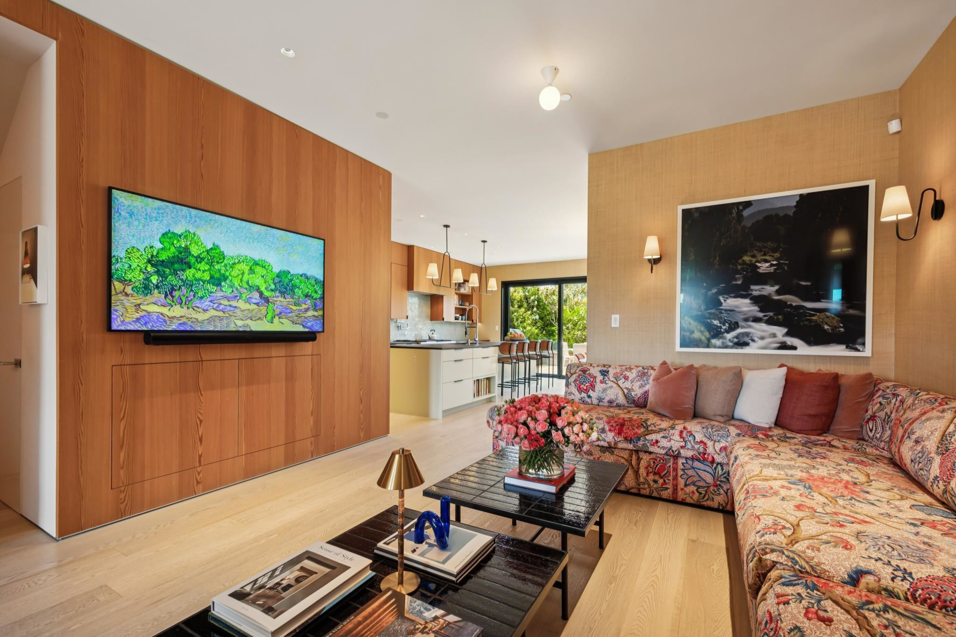 A bright living room with a floral-patterned sectional, black coffee tables with books and flowers, a wall-mounted TV, and a kitchen in the background.