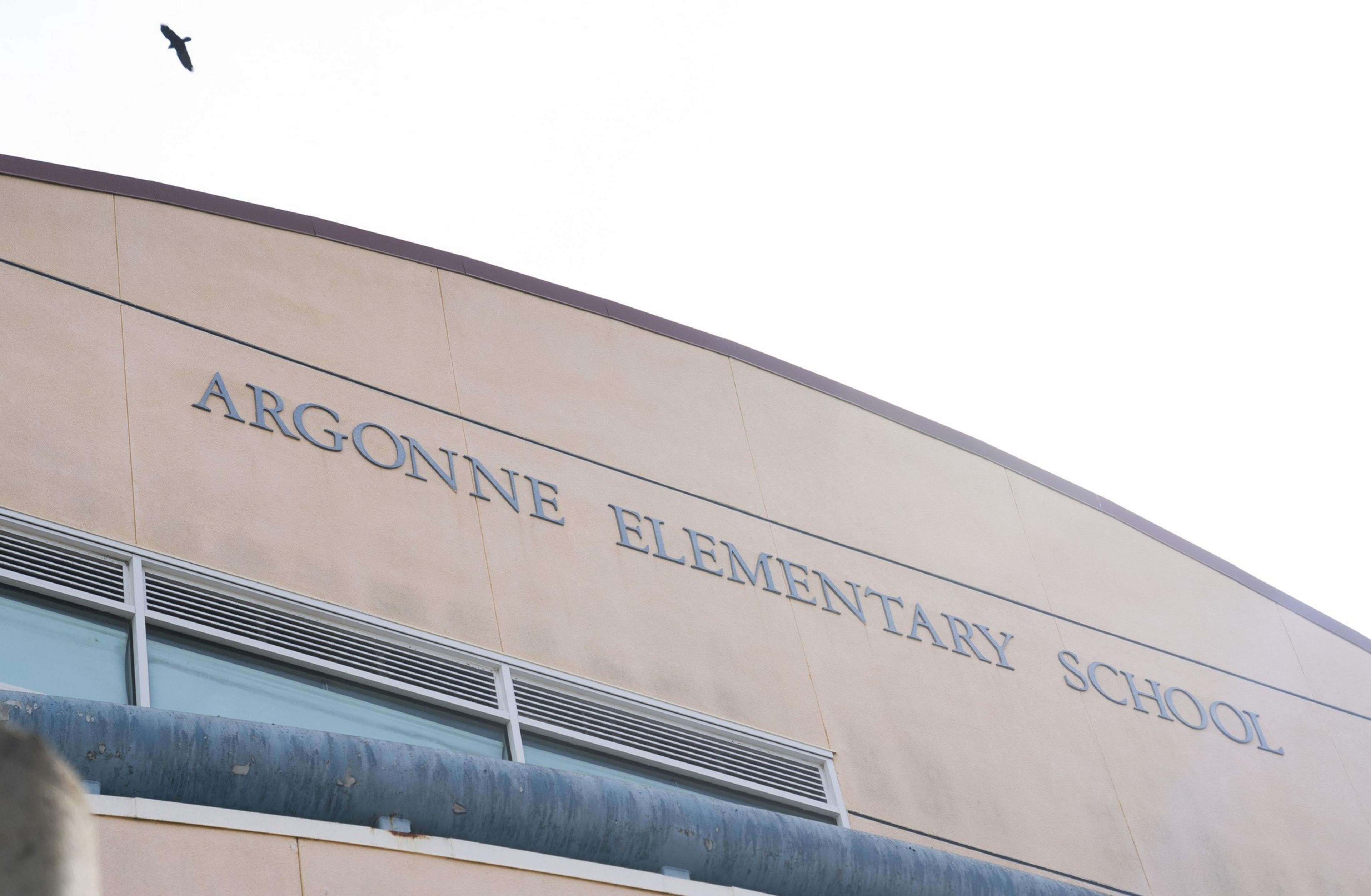 The image shows the beige facade of Argonne Elementary School with its name on the wall. A black bird flies above against a pale sky.
