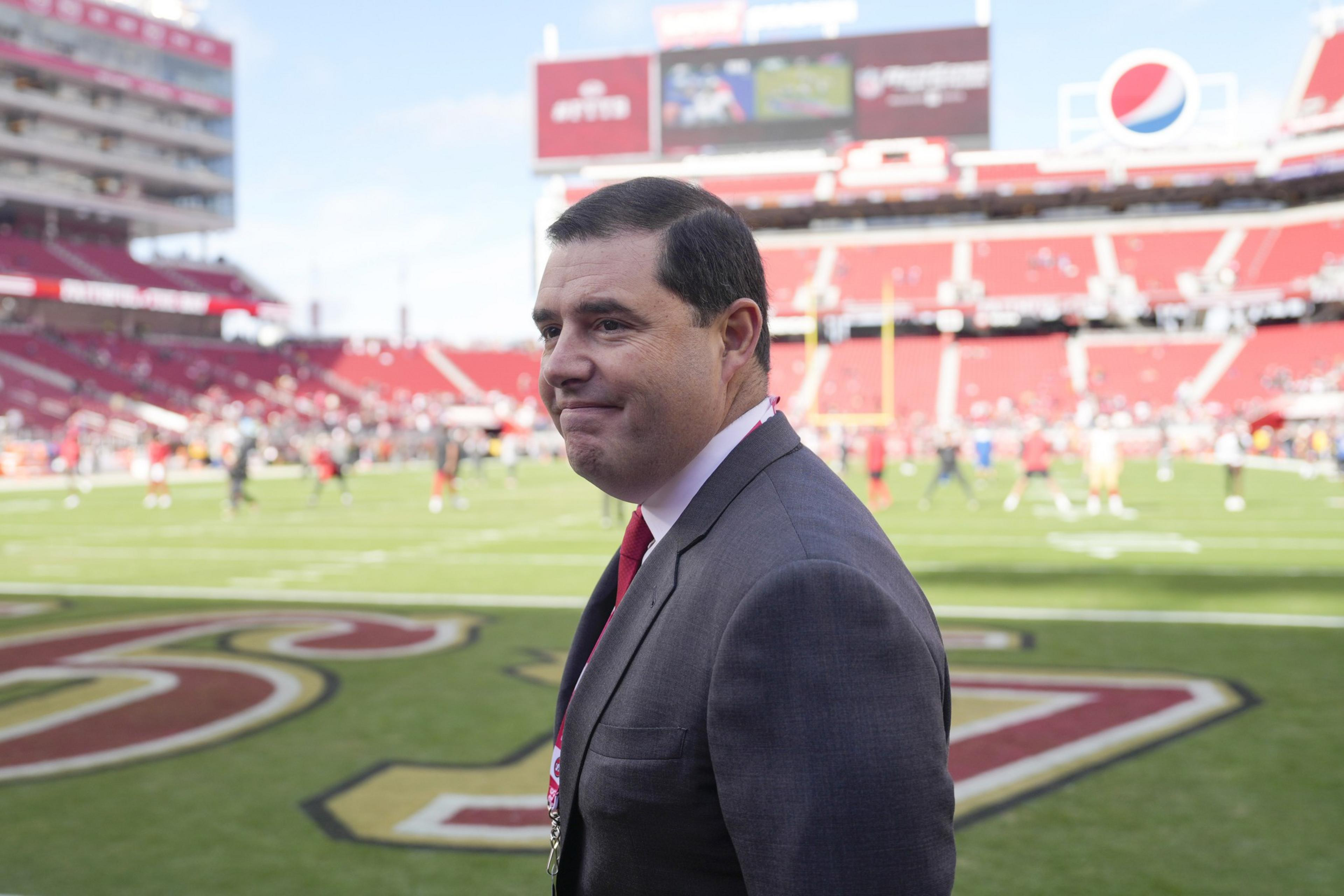 A man in a suit stands on a football field with red stadium seats and a scoreboard in the background. The field is marked with a large red and gold team logo.