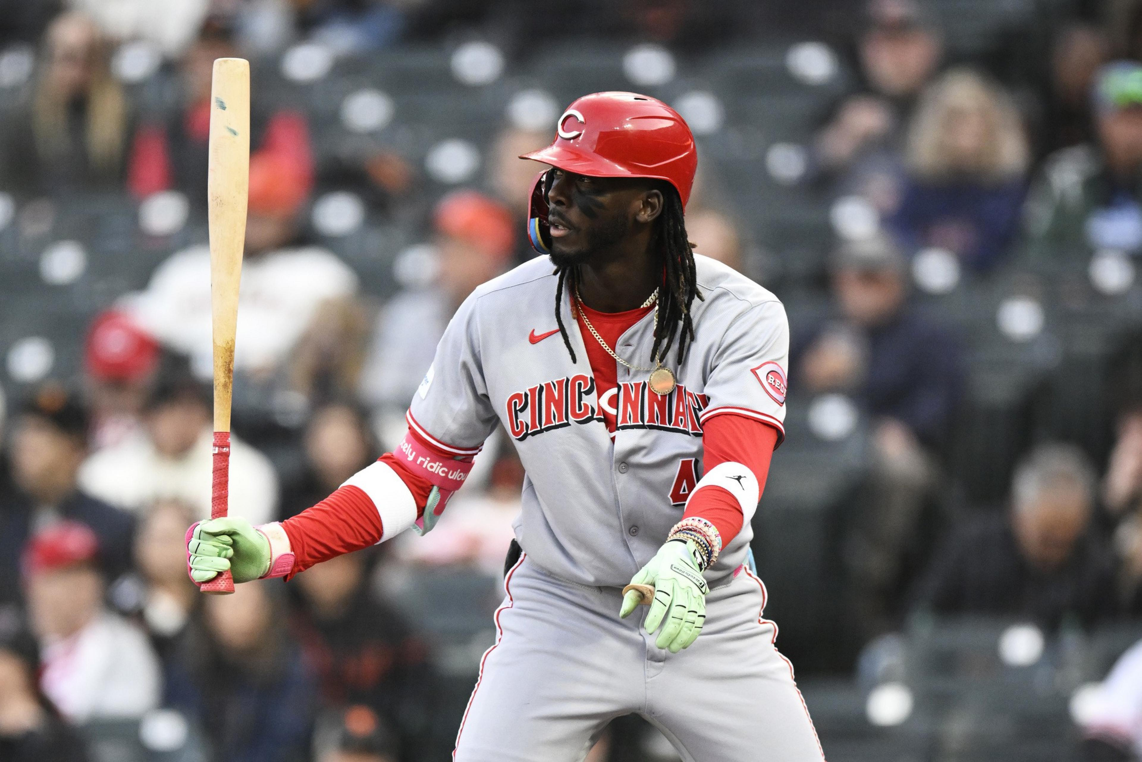 A baseball player in a Cincinnati uniform stands ready to bat. He's wearing a red helmet and green gloves, with the crowd blurred in the background.