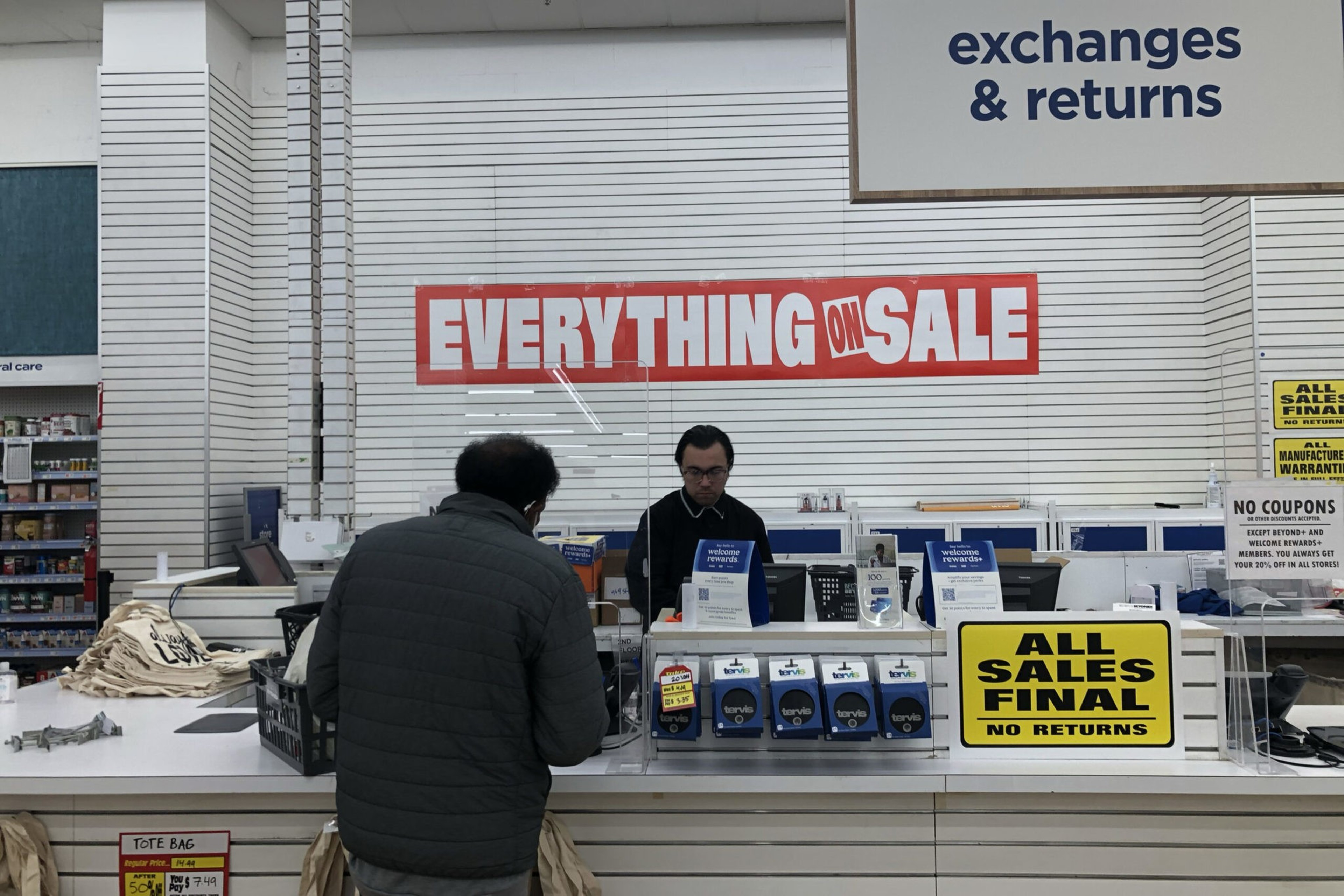 A man stands at a store counter beneath signs reading "EVERYTHING ON SALE," "ALL SALES FINAL NO RETURNS," and "exchanges & returns." A clerk is behind the counter.