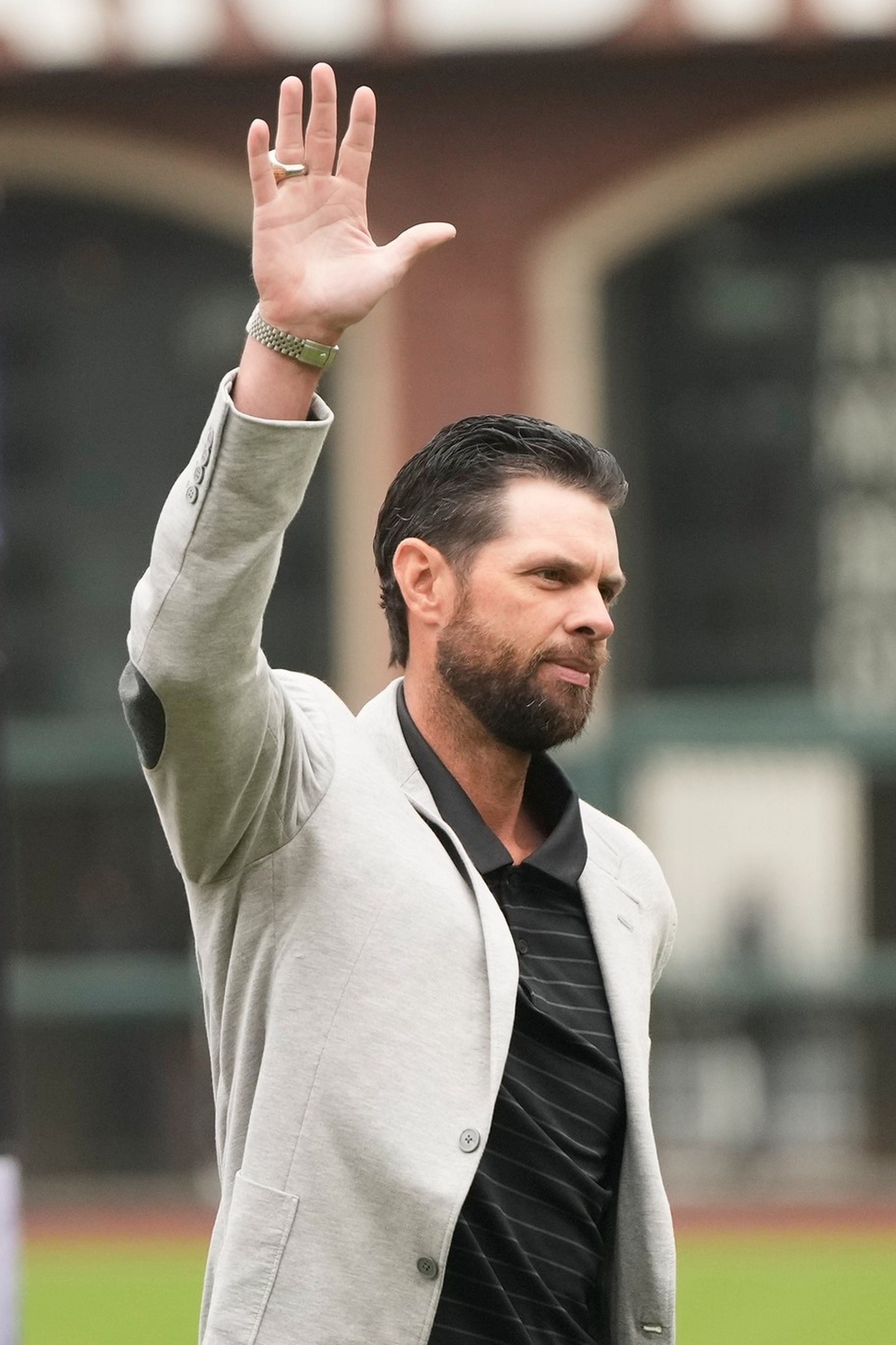 A man with a beard and slicked-back hair waves with his right hand ...