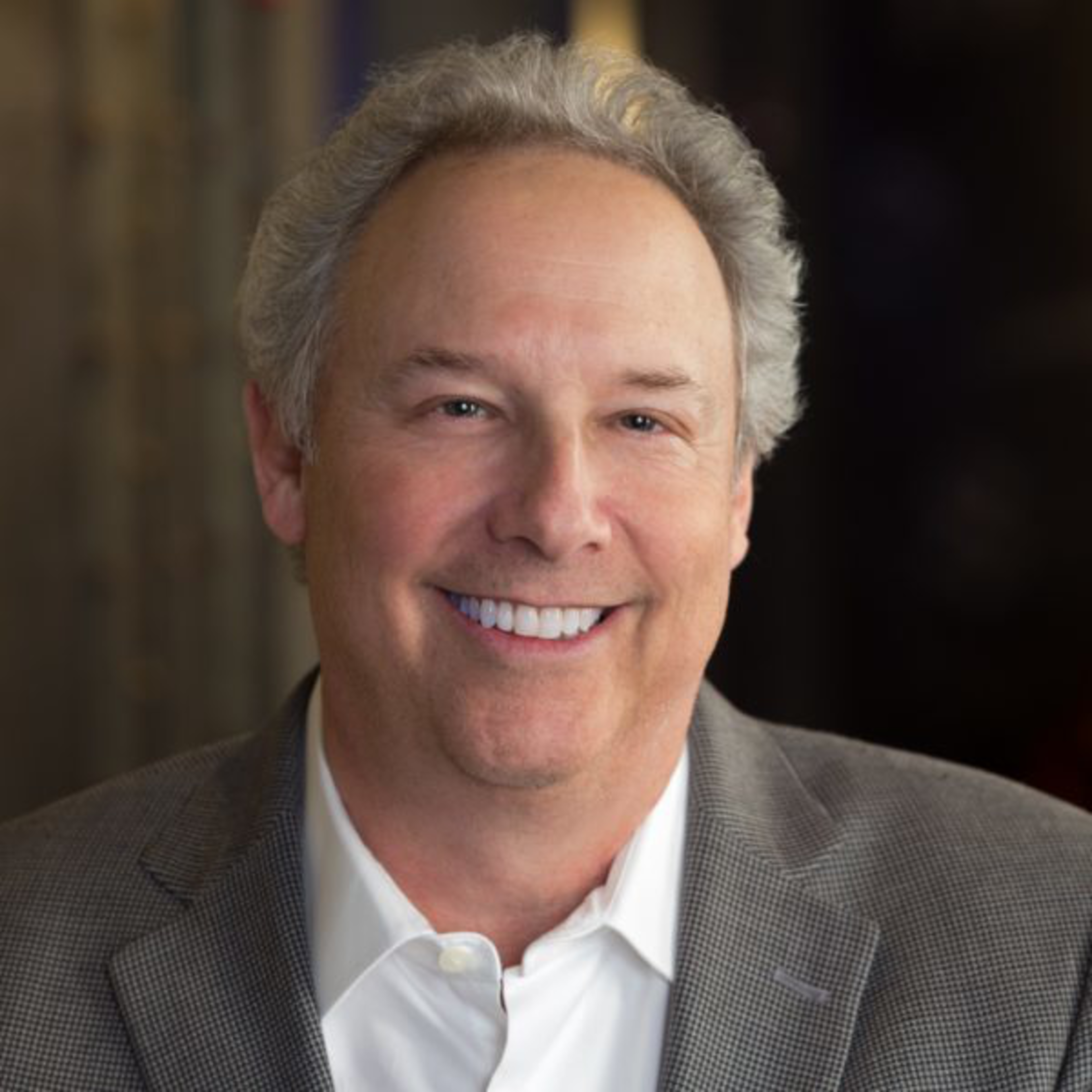 A middle-aged man with gray, slightly curly hair smiles warmly, wearing a white shirt and a gray blazer against a blurred dark background.