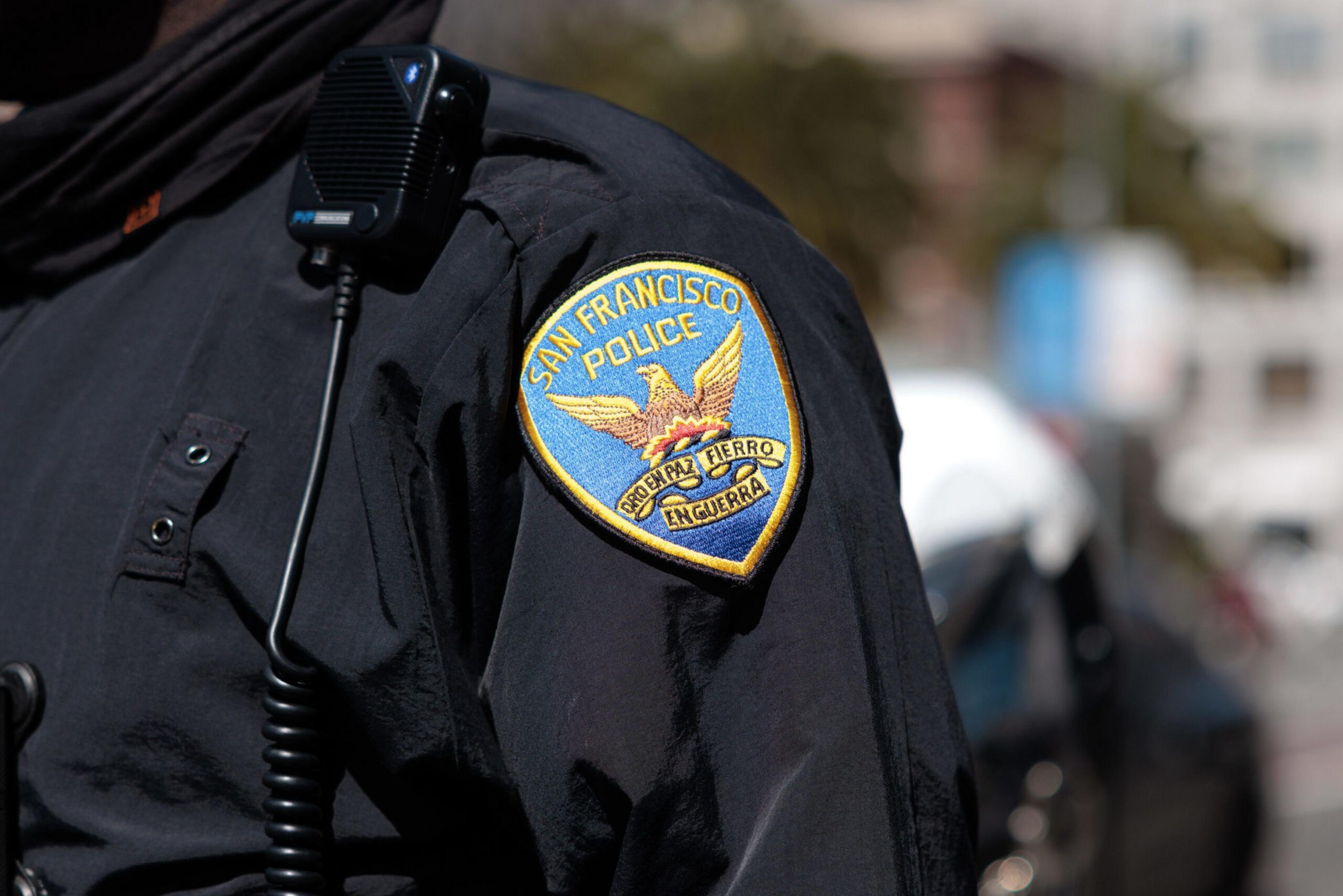 A San Francisco Police patch is visible on a dark uniform sleeve, accompanied by a shoulder microphone and blurred outdoor background.